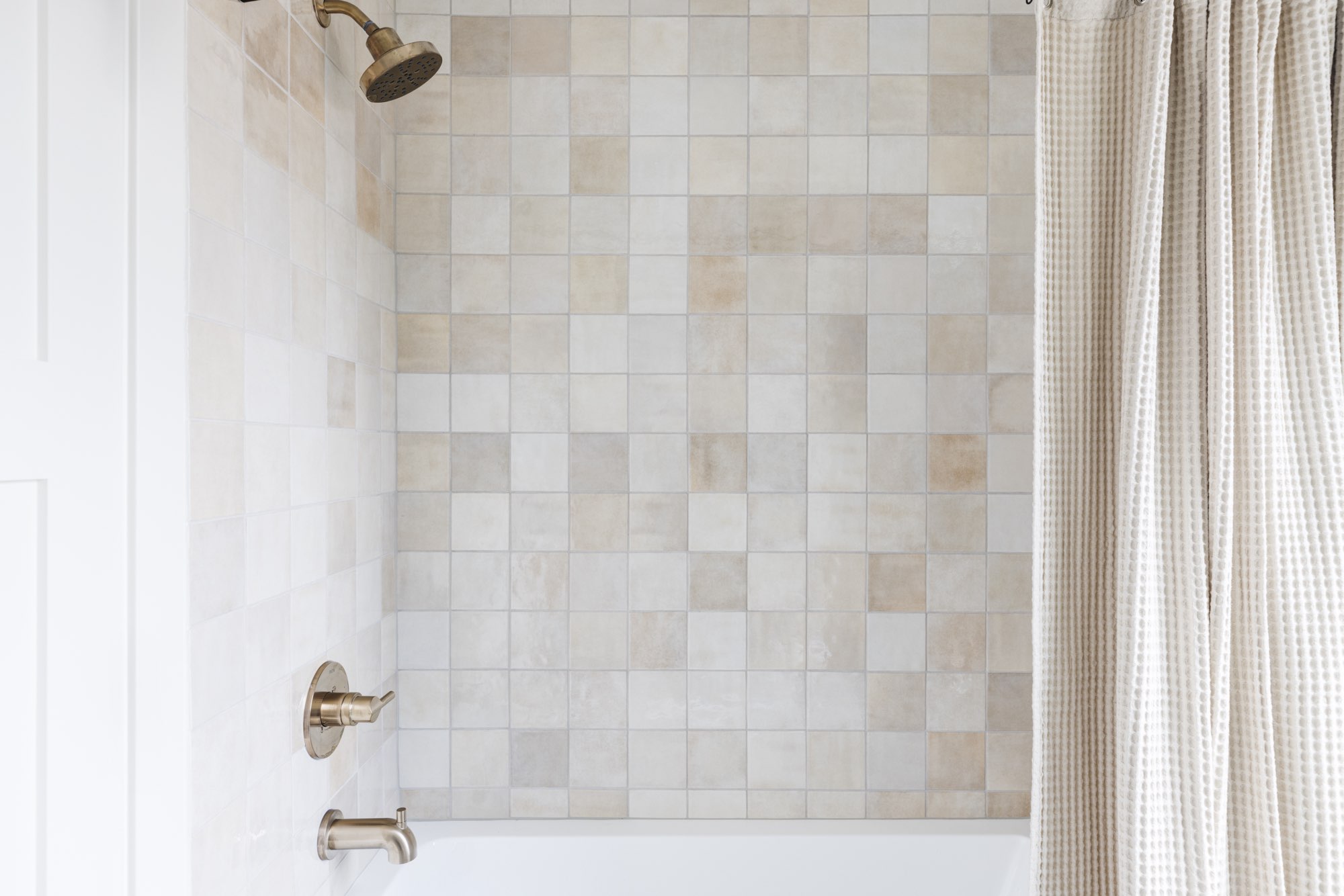 Tub and shower tiled in warm cream squares with a brass shower head and a striped curtain