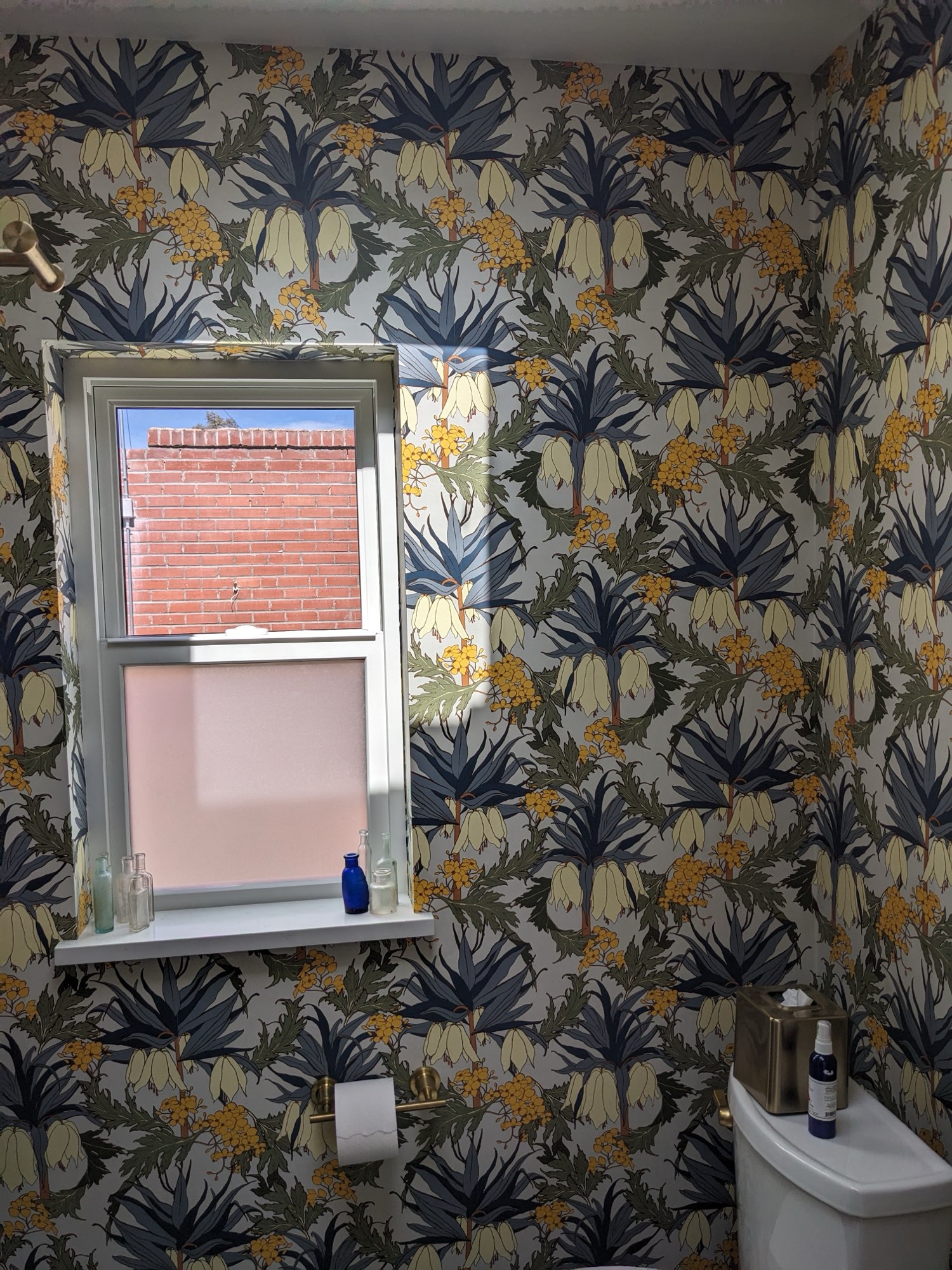 Powder room window with white trim and a frosted lower pane, surrounded by navy and yellow botanical wallpaper, with a brass toilet paper holder