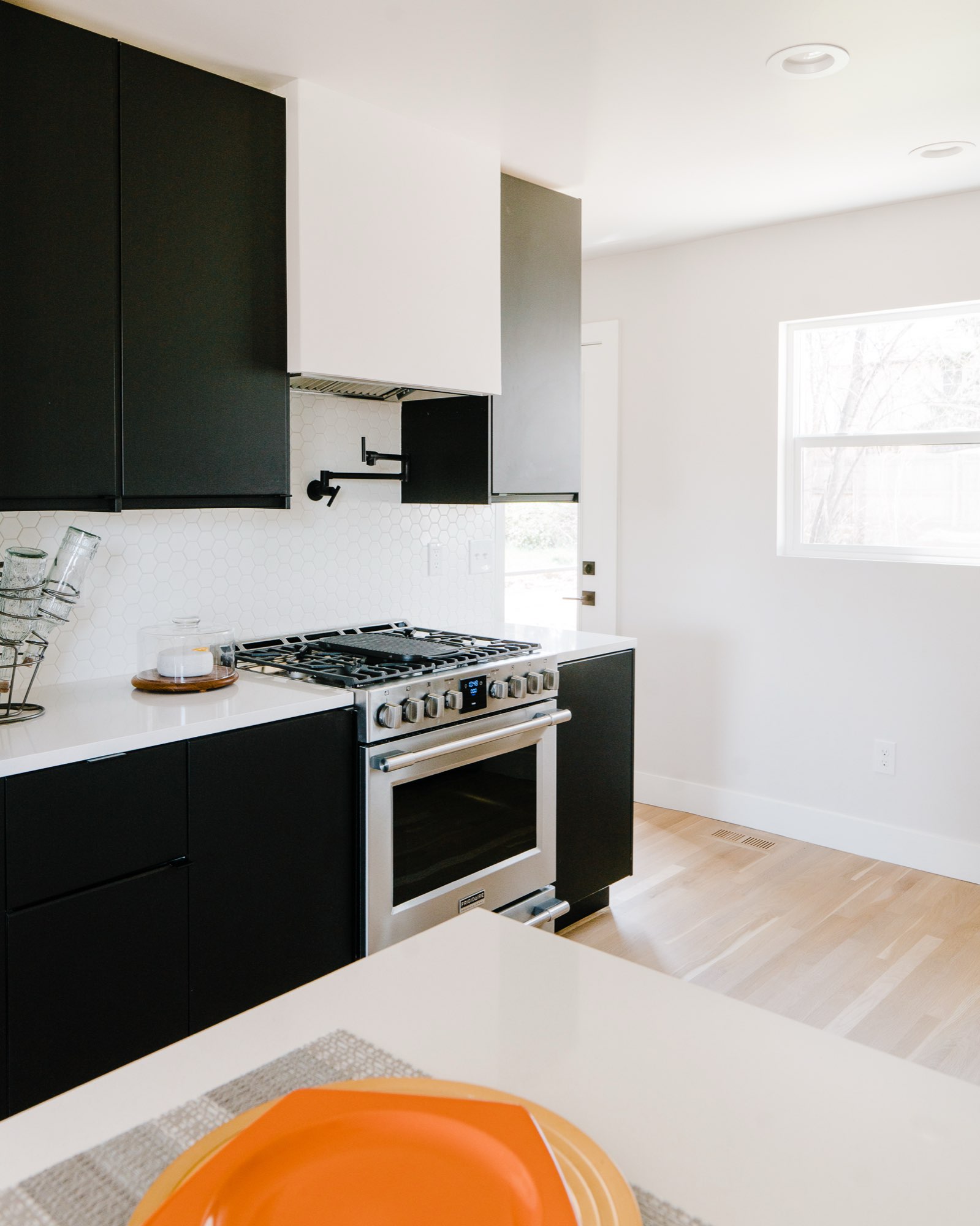 Black lower cabinets, white hood and stainless range beside a window in the bungalow kitchen