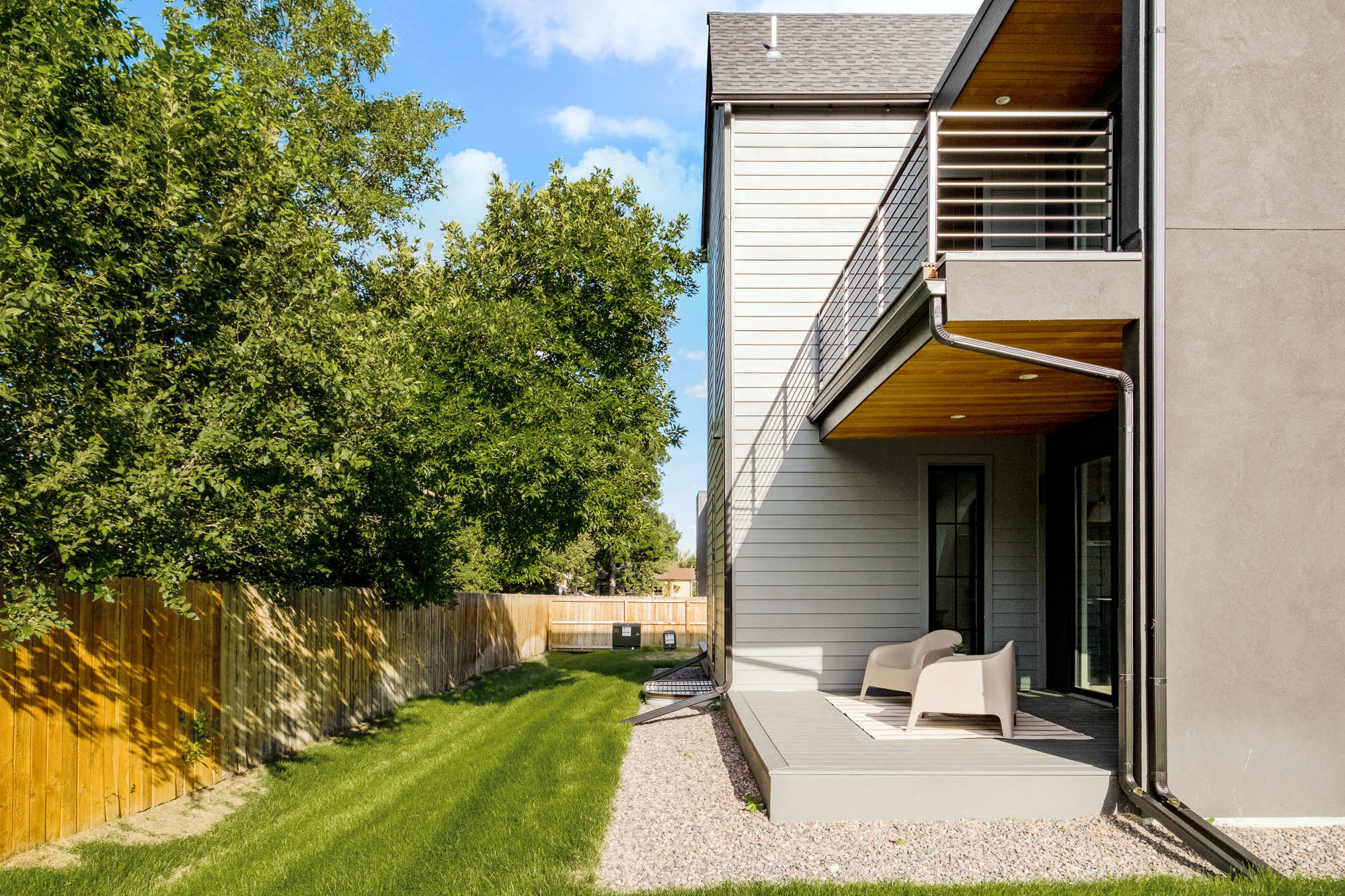 Side patio under the balcony, with a gravel strip, gray siding and a fenced yard
