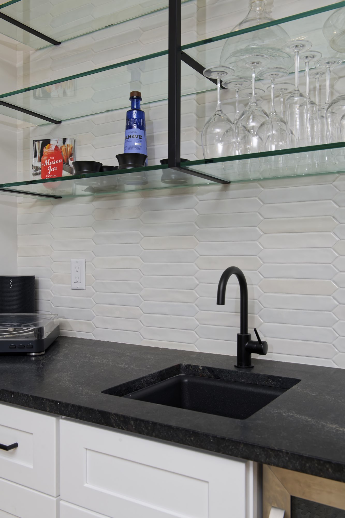 Wet bar sink with a black faucet and glass shelves over a white tile backsplash