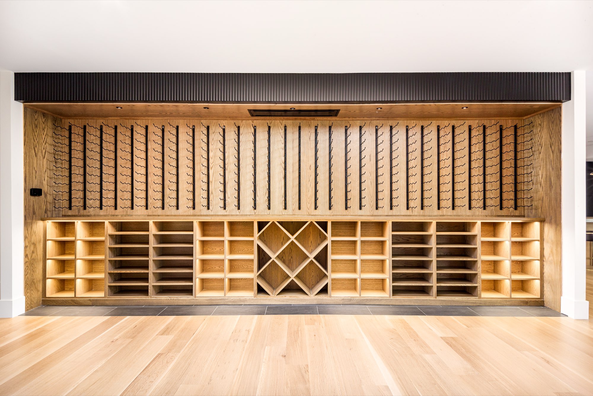 Full wall of wood shelving, black metal wine racks and lit cubbies above light wood floors