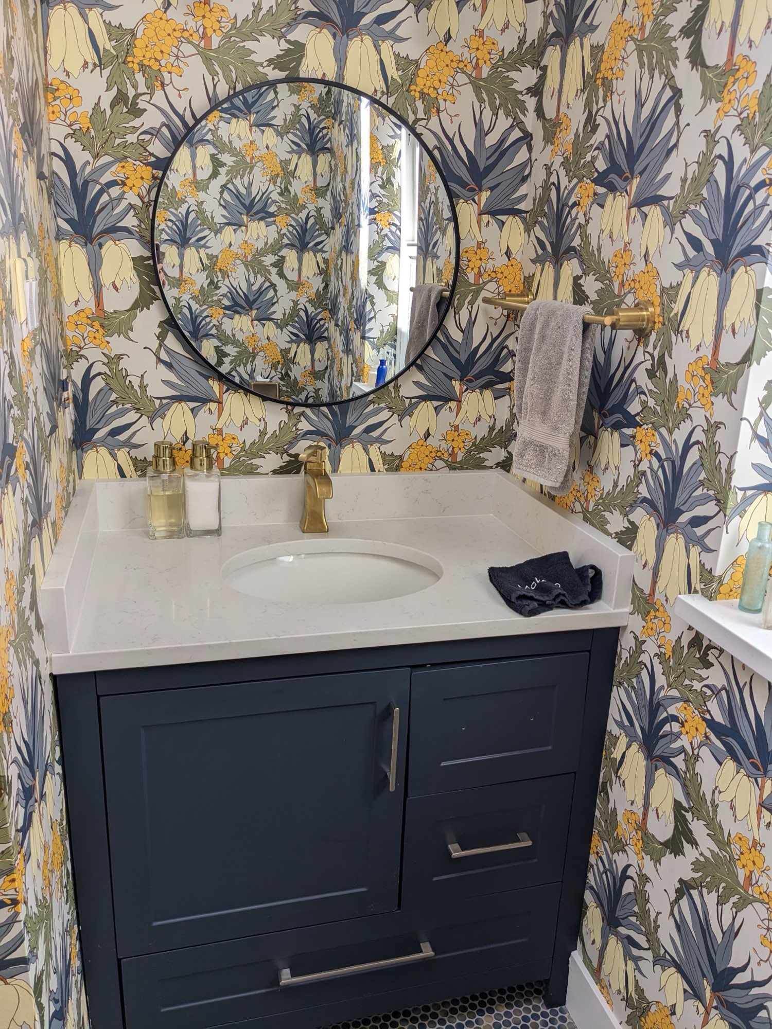 Powder room with a navy vanity, white stone top, round black mirror, brushed brass faucet and towel bar, and botanical wallpaper in navy, yellow and cream