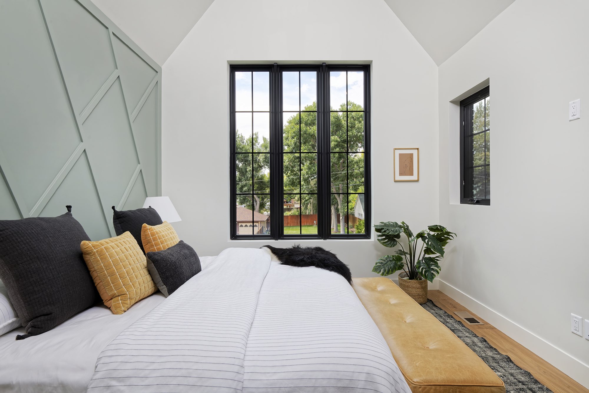 Bedroom corner with the sage paneled wall, a black-framed window and a leather bench at the foot of the bed