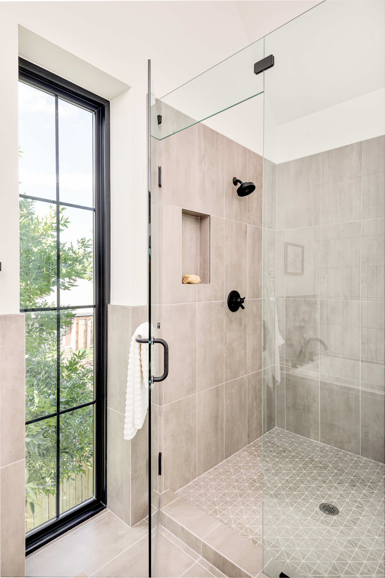 Glass shower in warm gray tile with a niche, a black shower head and a tall window