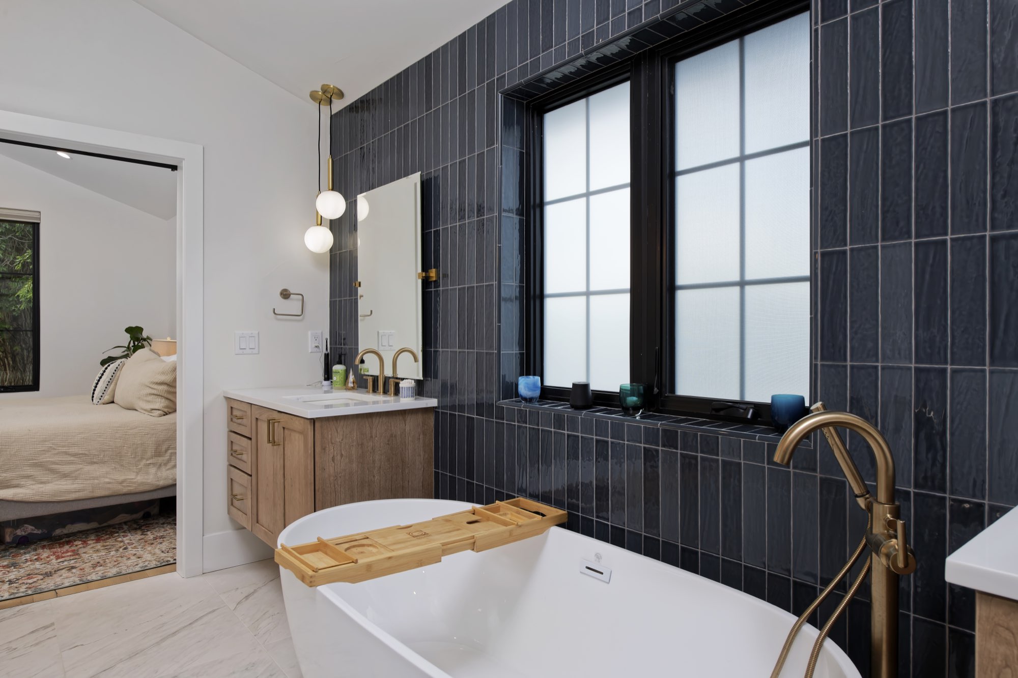 Primary bath with navy tile, a freestanding white tub, brass faucet and a wood vanity under black-framed windows
