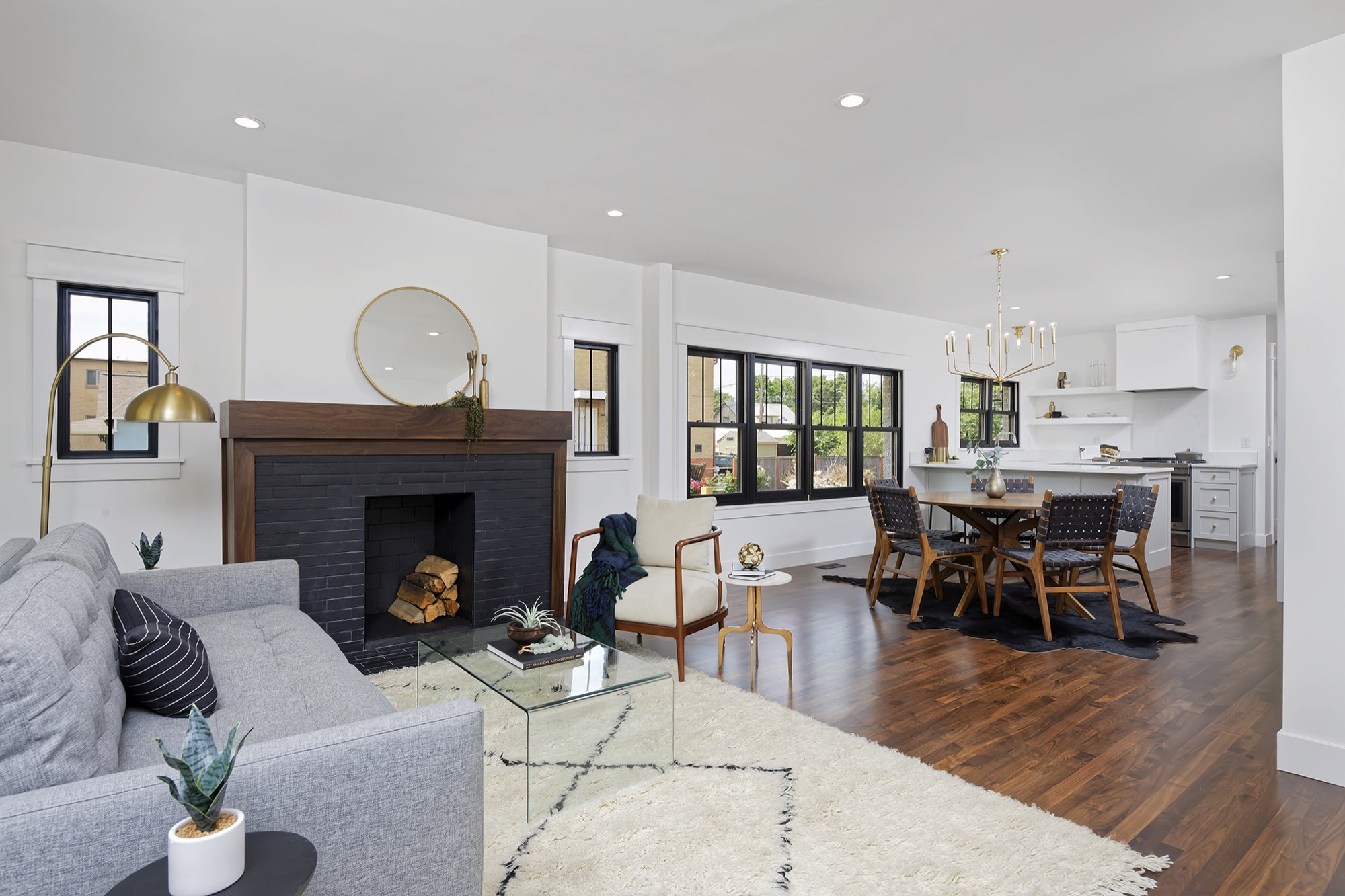 Open living, dining and kitchen seen from the fireplace, with a brass chandelier and a row of black-framed windows