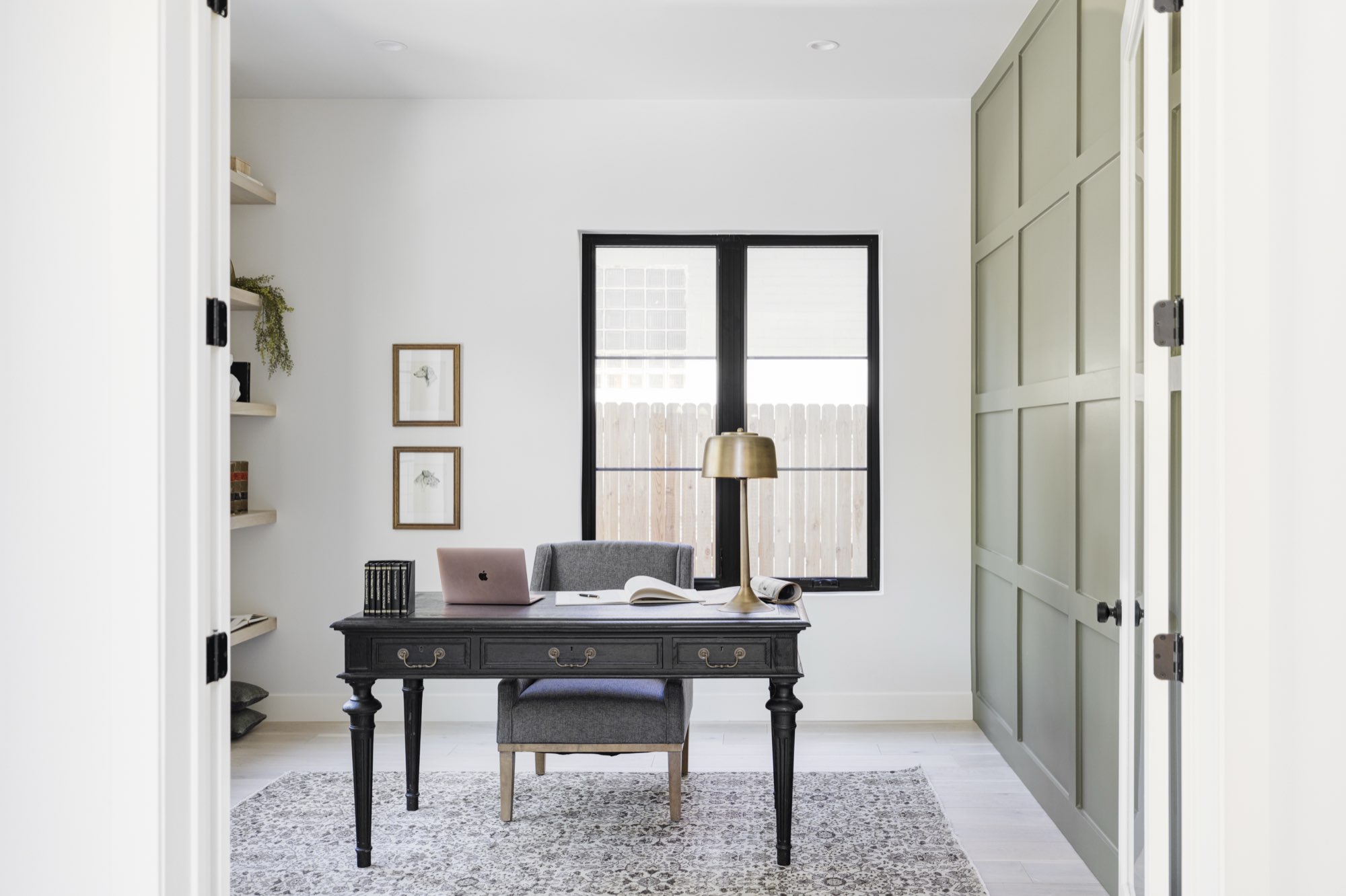 Home office with a black-framed window, open shelves and a wall of sage green paneling seen through double doors