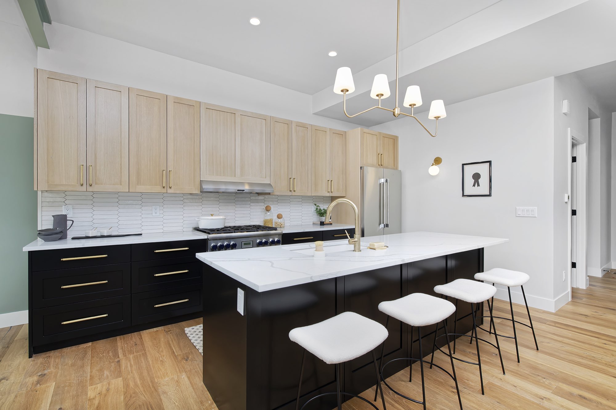 Wide view of the kitchen with light wood cabinets, a black island with white counter, brass chandelier and white stools