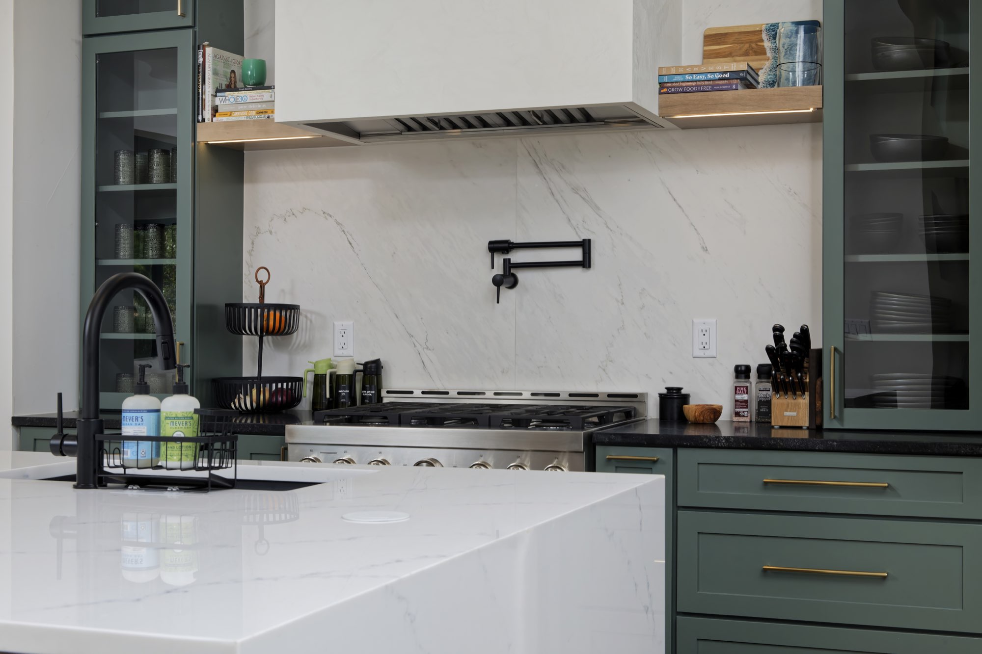 Island corner with a black faucet, sage green cabinets with glass doors and a white slab backsplash