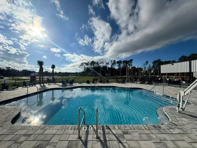 The resort pool at Heritage Oaks RV Resort under a partly cloudy sky, framed by palms and loungers