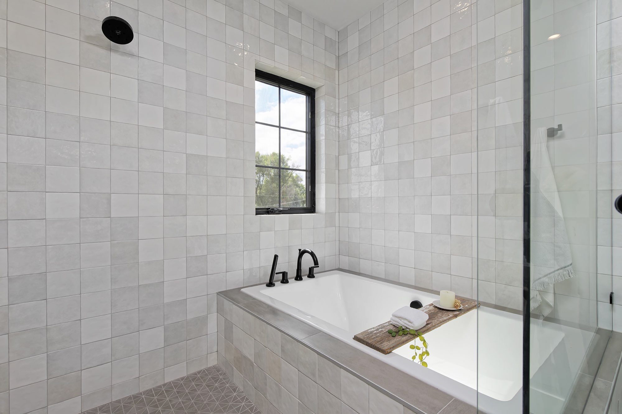 Soaking tub set into a tiled deck with black faucets, glass shower panel and a window in the soft gray tile wall