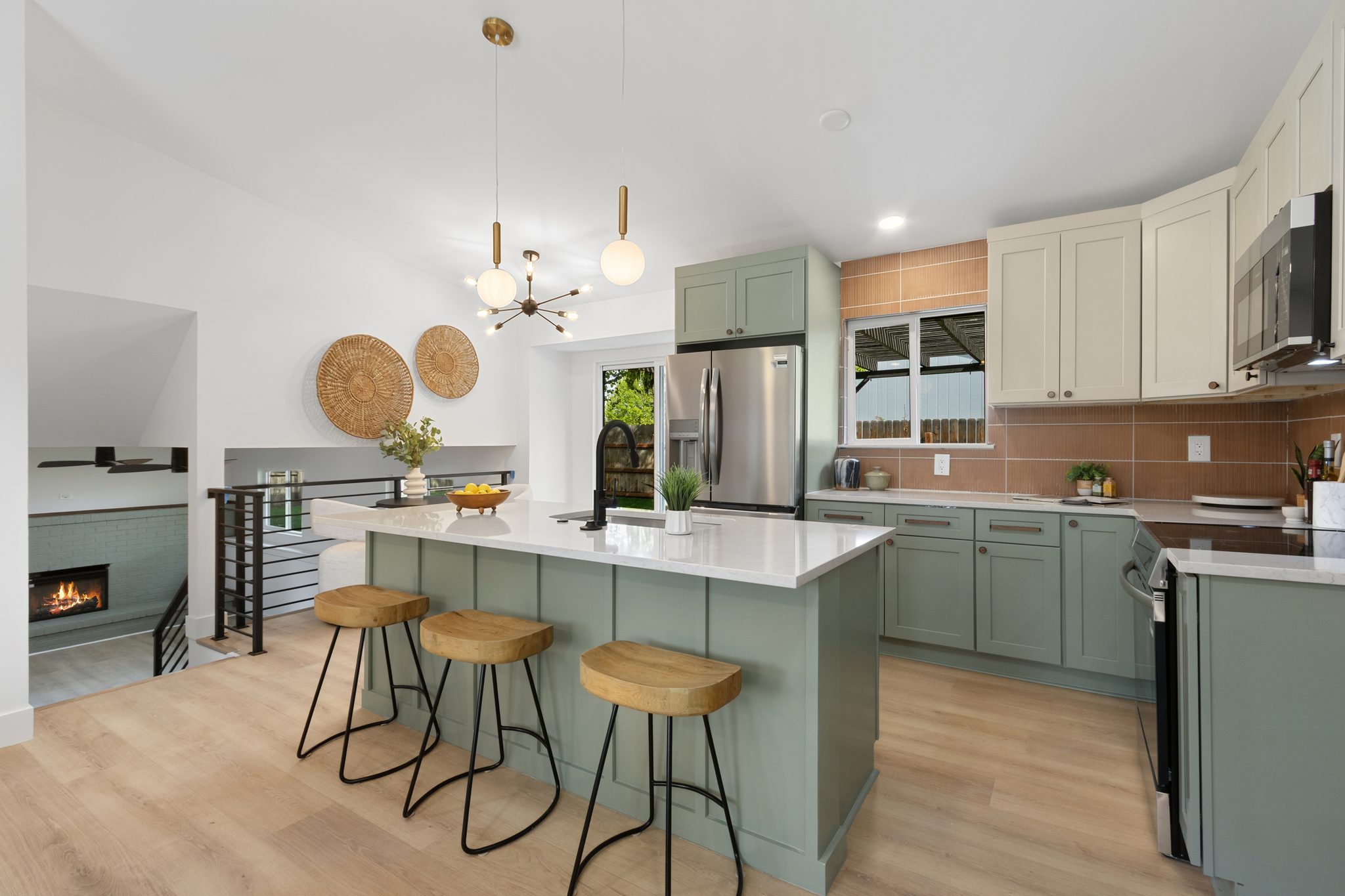Wide view of the kitchen with the sage island and three stools, the range wall, the refrigerator and a view toward the lower-level fireplace