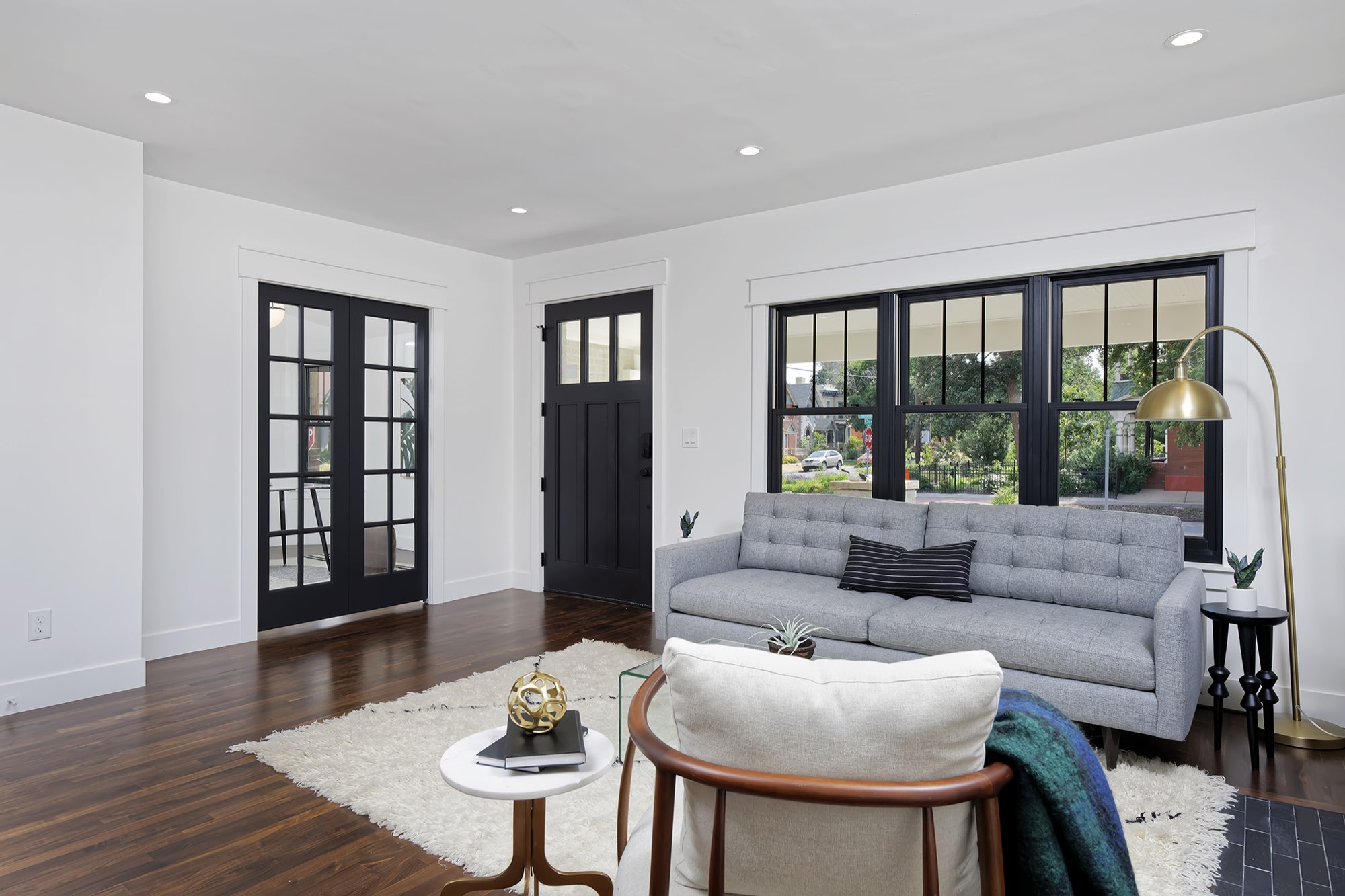 Living room with dark wood floors, a gray sofa, black French doors, a black front door and a row of black-framed windows