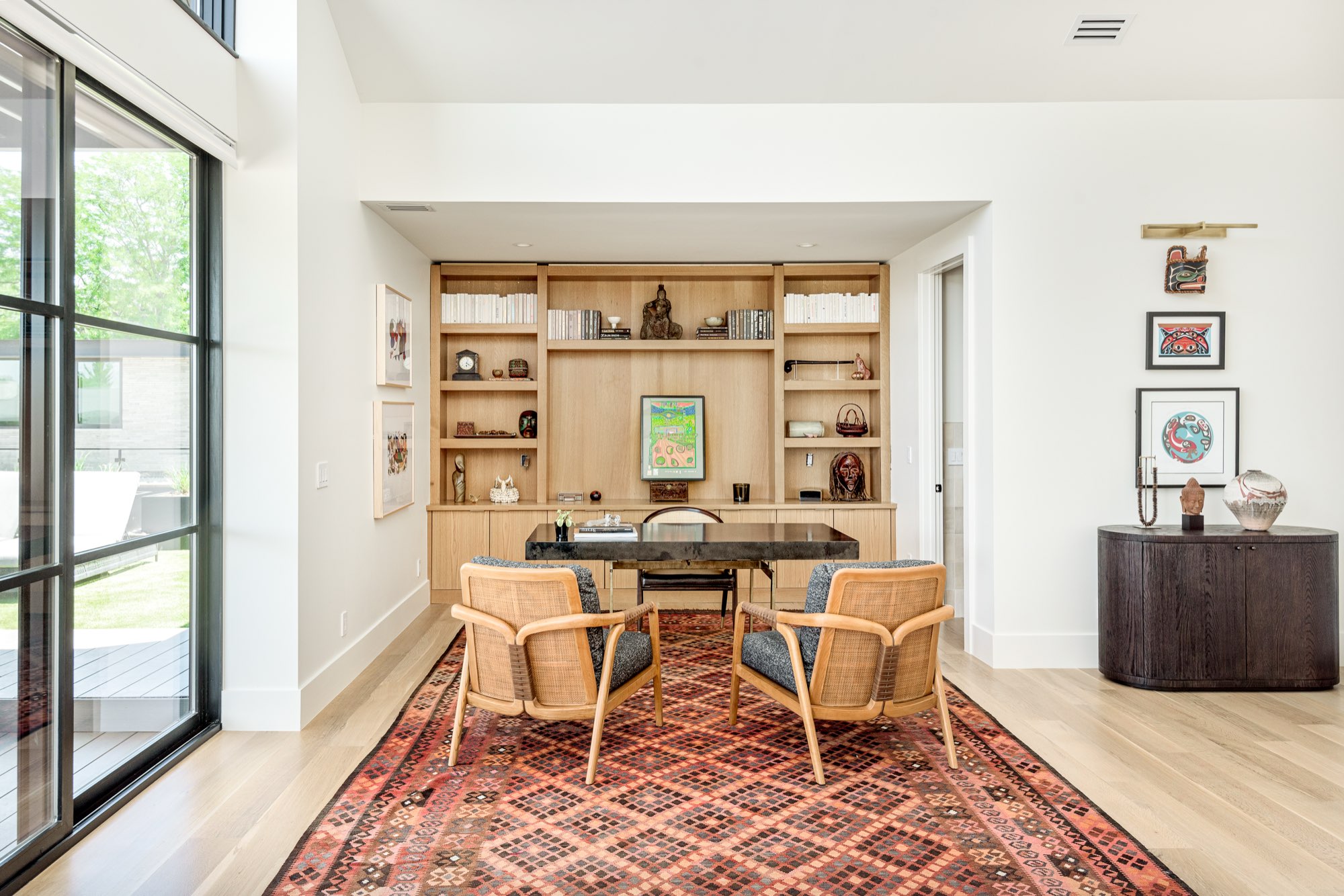 Study with built-in light wood shelving, a dark desk, two woven chairs and black-framed glass doors