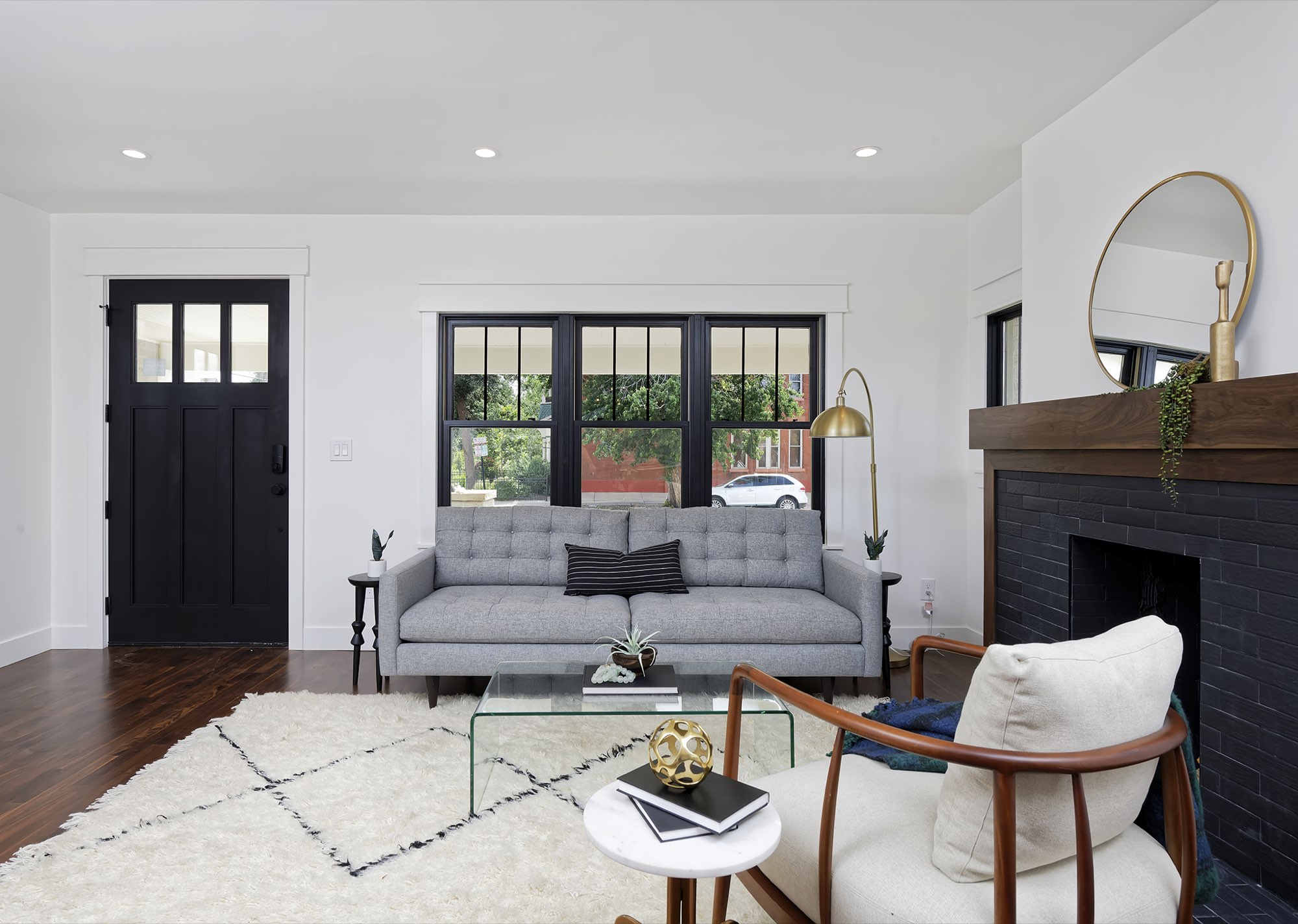 Living room with a black front door, gray sofa, rug and fireplace beside a wall of black-framed windows
