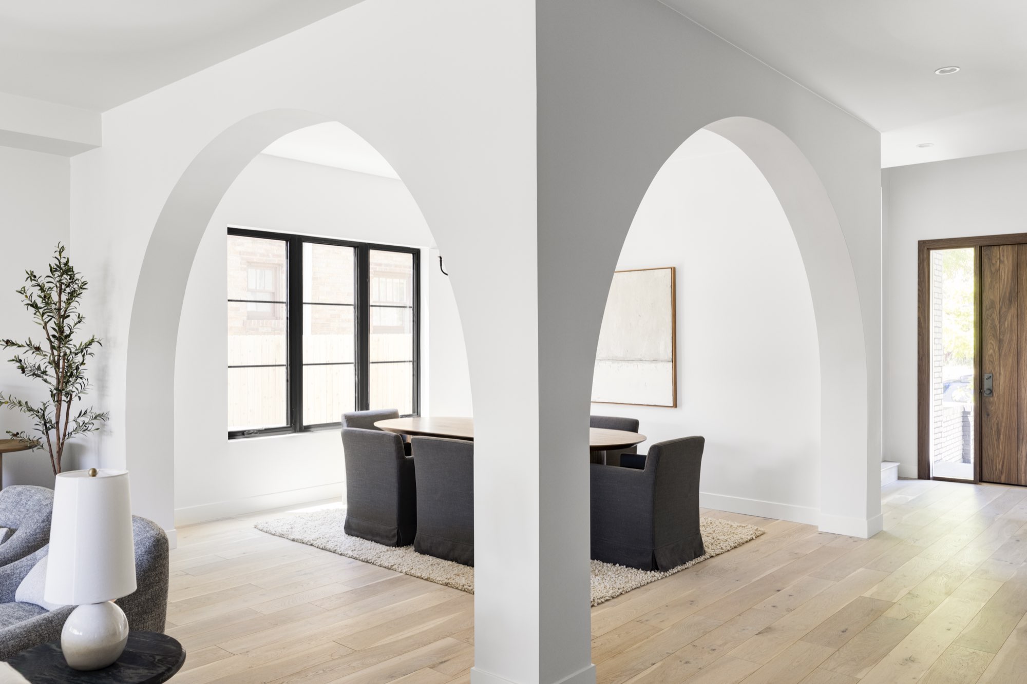 Two tall arched openings framing a dining area with black-framed windows and light wood floors