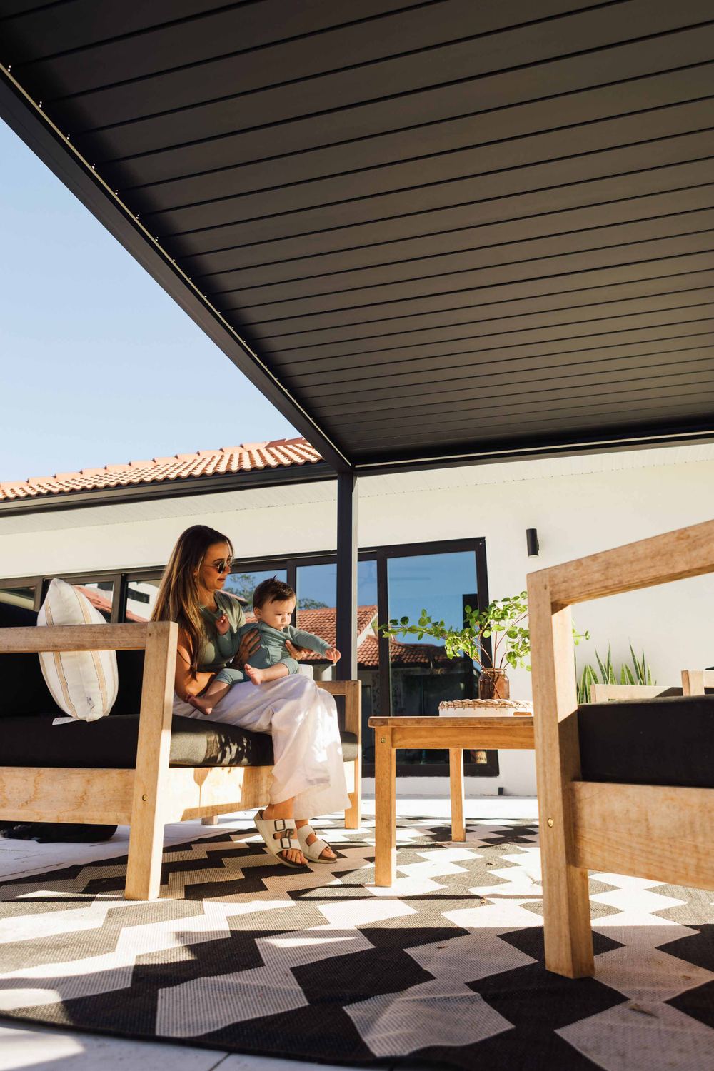 Family relaxing under a dark louvered PERGOLUX roof on a sunny patio