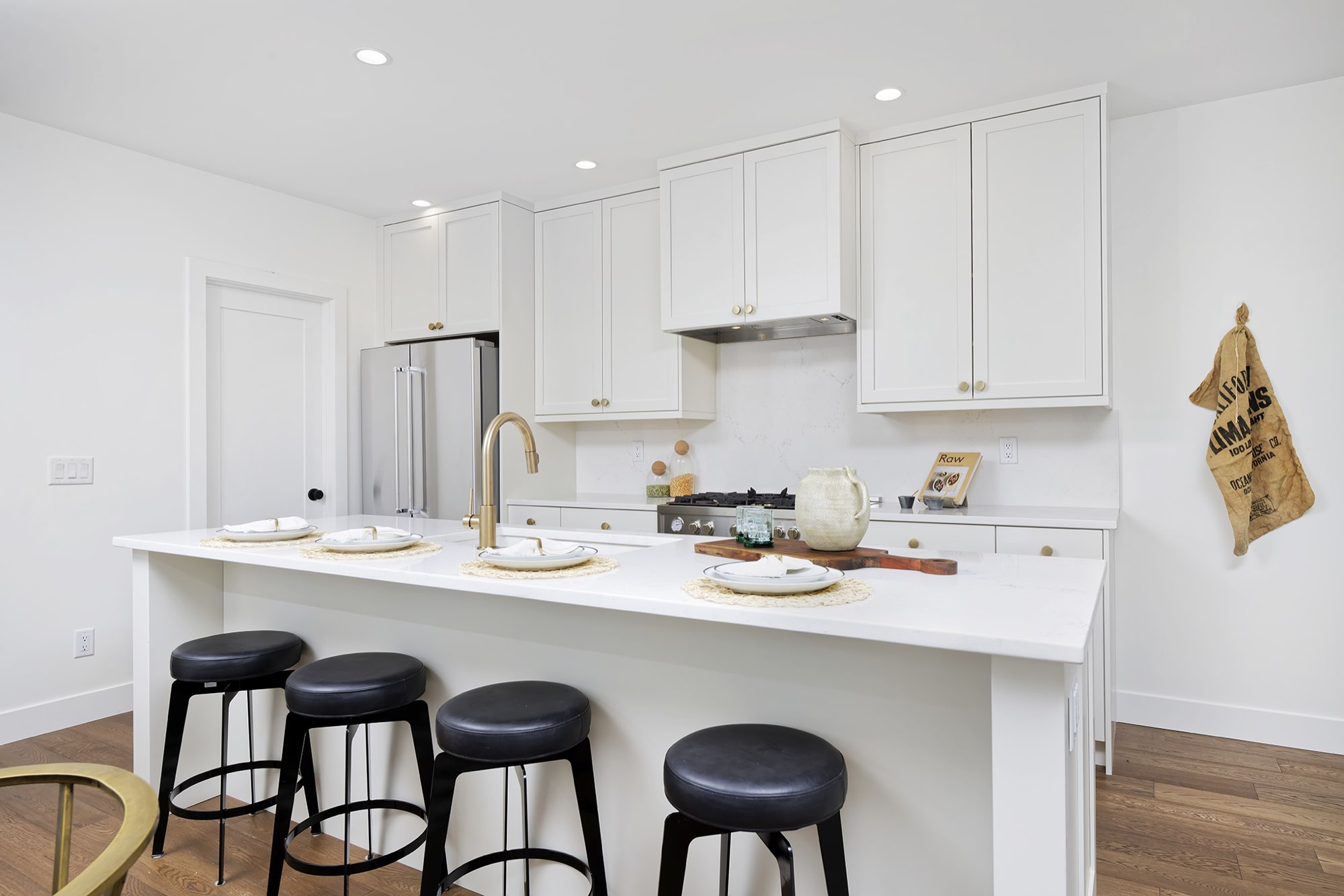 Wide view of the white kitchen with a long island, four black stools, shaker cabinets and a stainless refrigerator