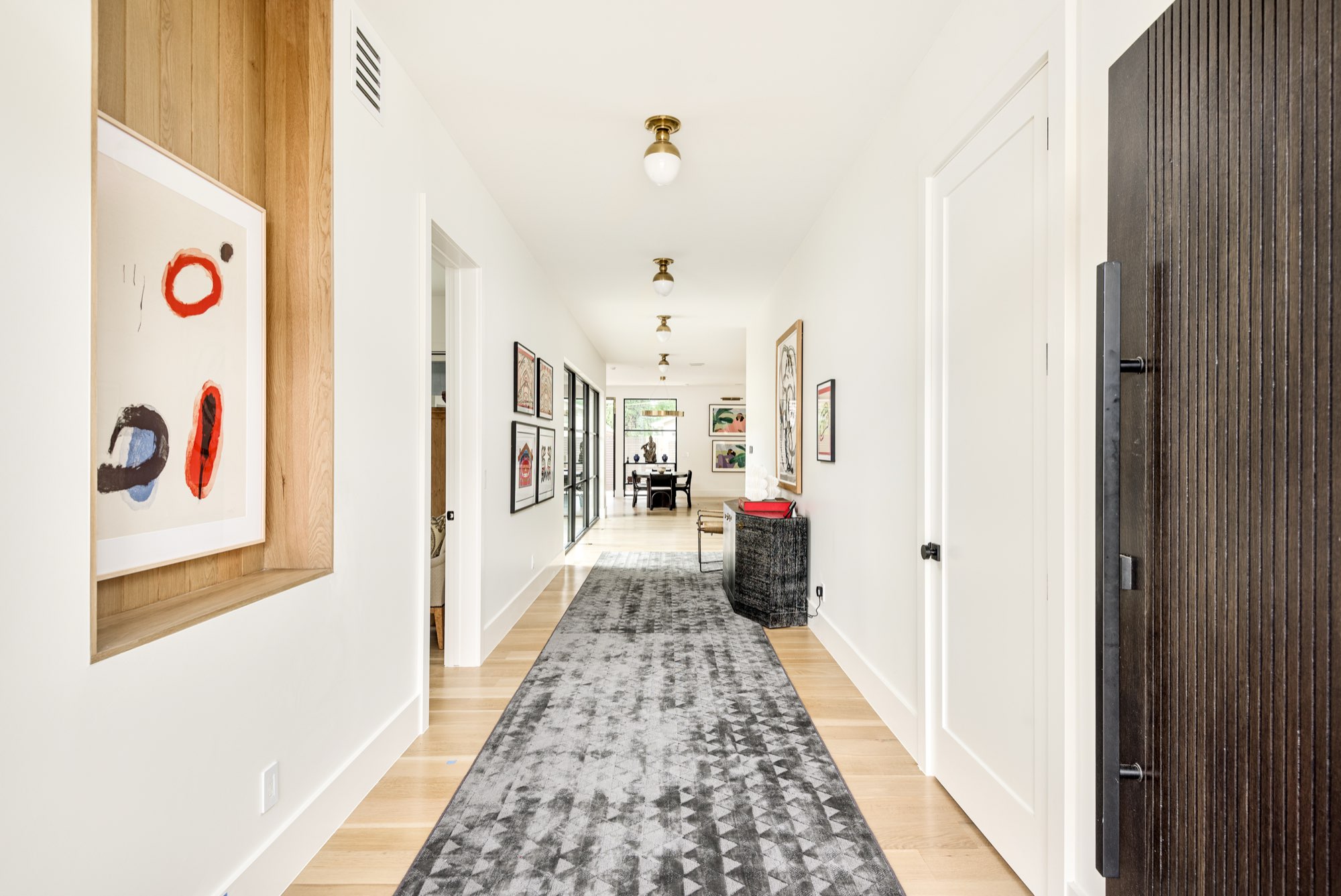 Long hallway with light wood floors, a dark wood door and framed art, with a dining room at the far end