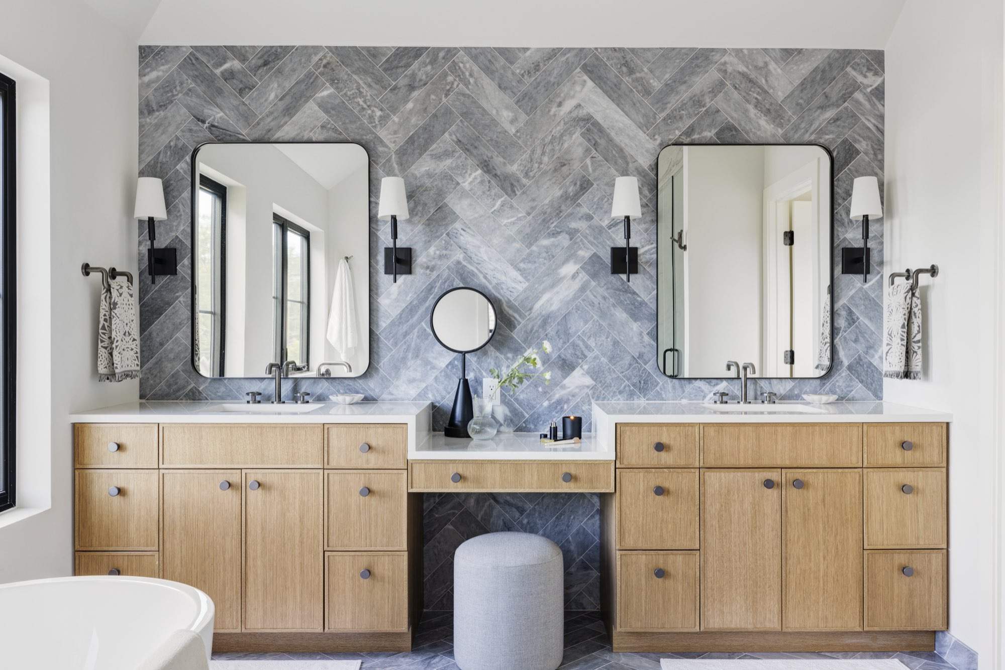 Double vanity with light wood cabinets, two black-framed mirrors, black sconces and a gray herringbone tile wall