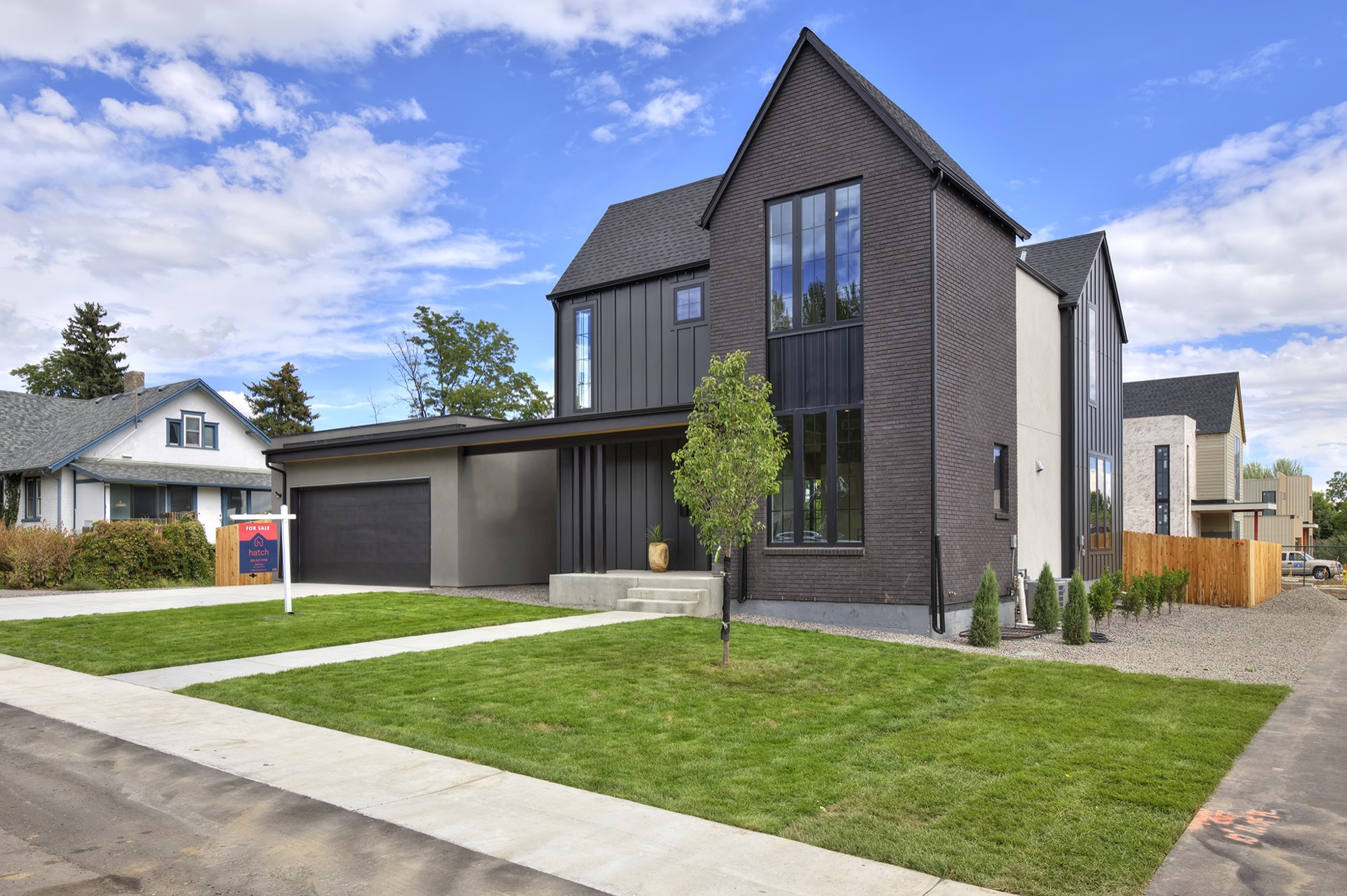 Angled front view of the modern farmhouse with a dark brick tower, tall black-framed windows, charcoal siding and a young tree in the lawn
