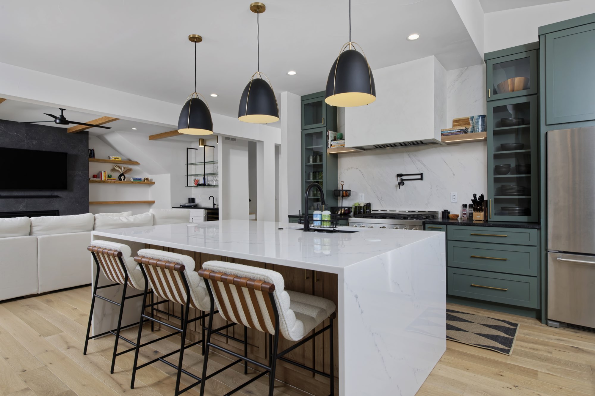 Wide kitchen with a long white waterfall island, black pendants, sage green cabinets and a white hood