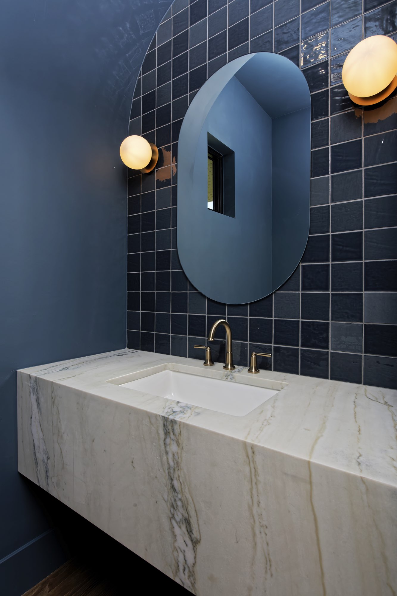 Powder room with blue square tile, an oval mirror, round brass sconces and a floating veined stone vanity