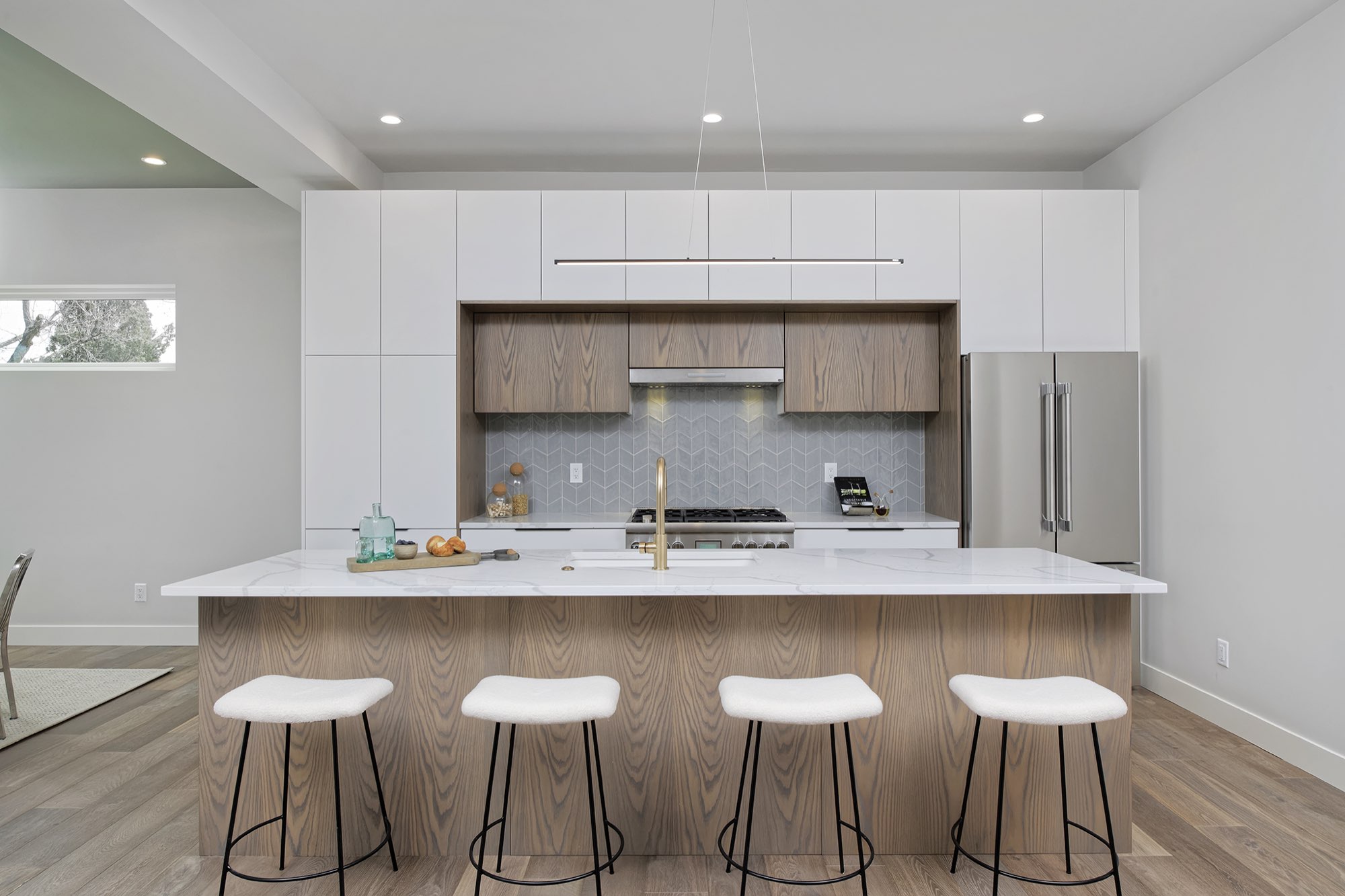Straight-on view of the kitchen with a long wood-grain island, four white stools, white upper cabinets and a chevron tile backsplash