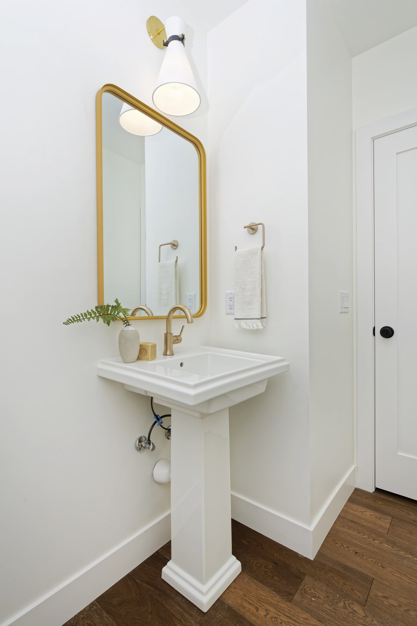 Powder room with a pedestal sink, brass-framed mirror and wall sconce above wood floors