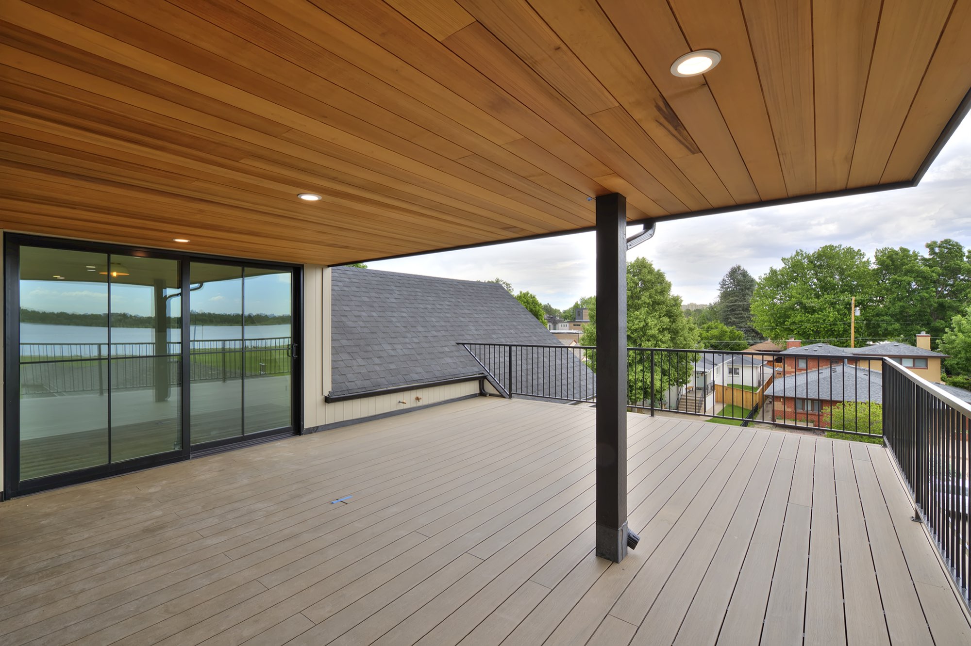 Covered deck seen from under the wood-plank ceiling, with sliders on the left and a black railing over the neighborhood