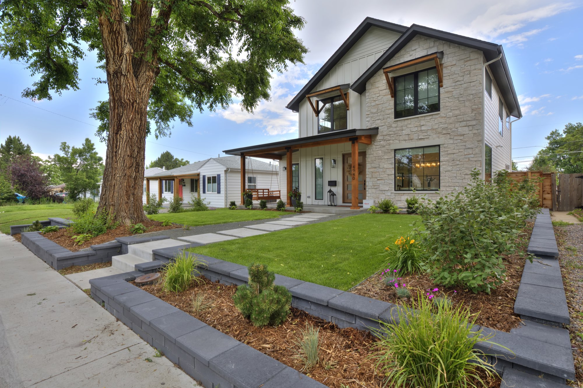 Front of the new custom home in Denver with white board-and-batten siding, light stone, a timber-post covered porch and a landscaped front yard