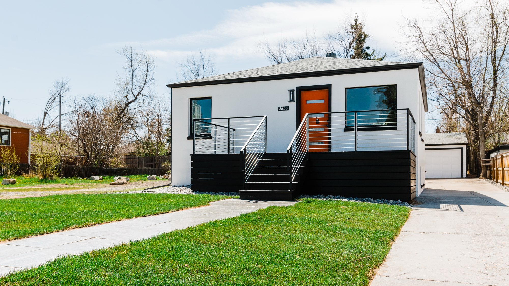 Front of a renovated 1920s bungalow in Denver with white walls, black trim, an orange front door and a dark deck with a cable railing