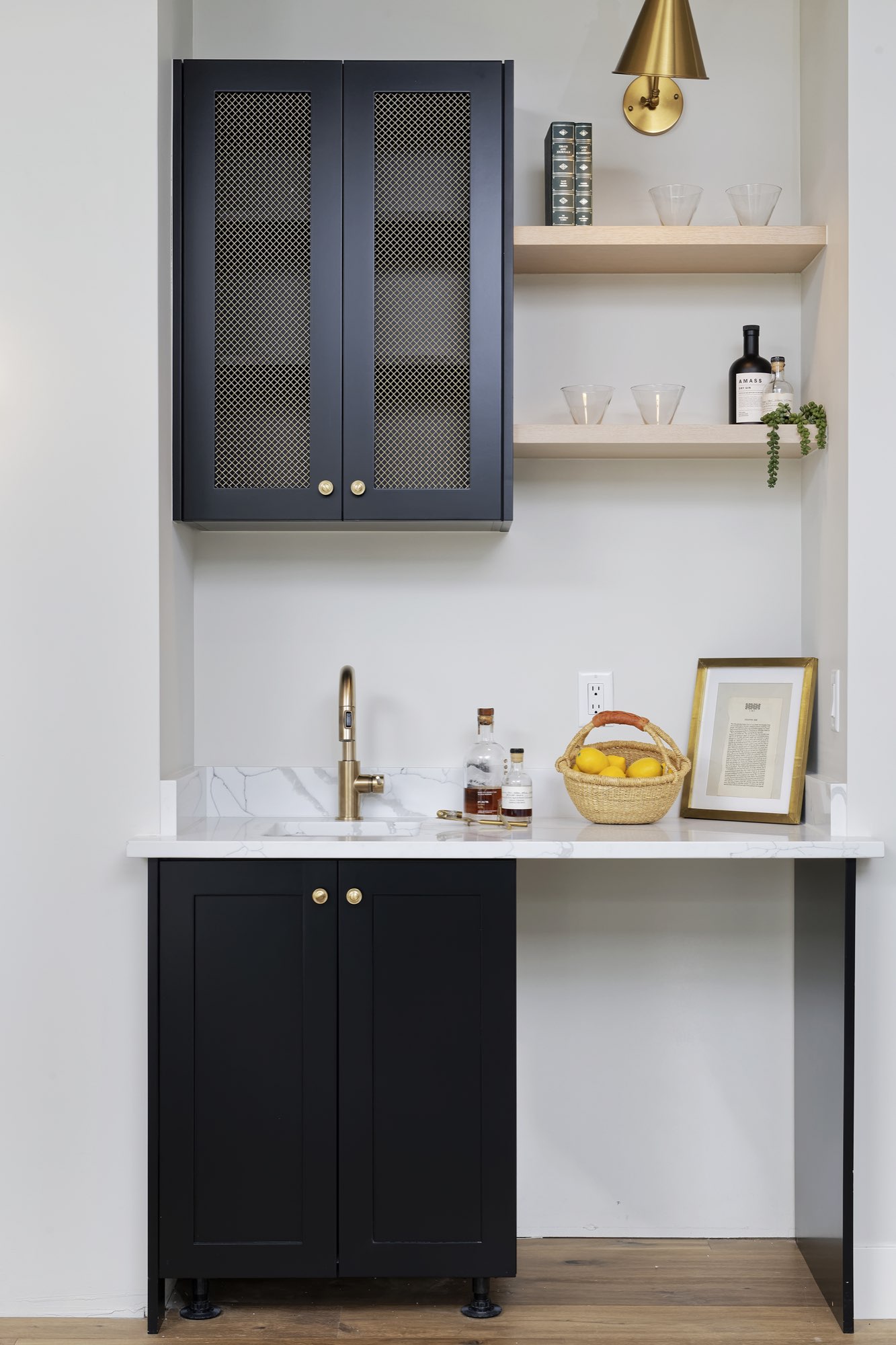 Wet bar niche with black cabinets, mesh-front upper doors, floating wood shelves, brass faucet and brass sconce
