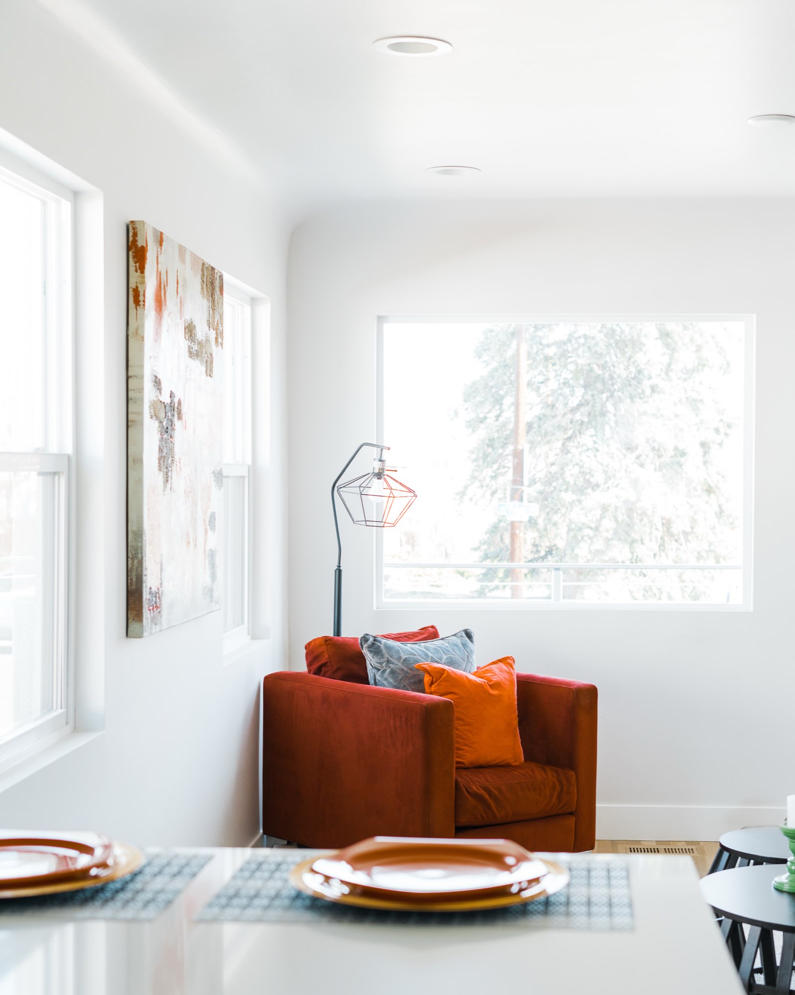 Rust armchair with orange pillows beside a window and an abstract painting in the corner of the living room