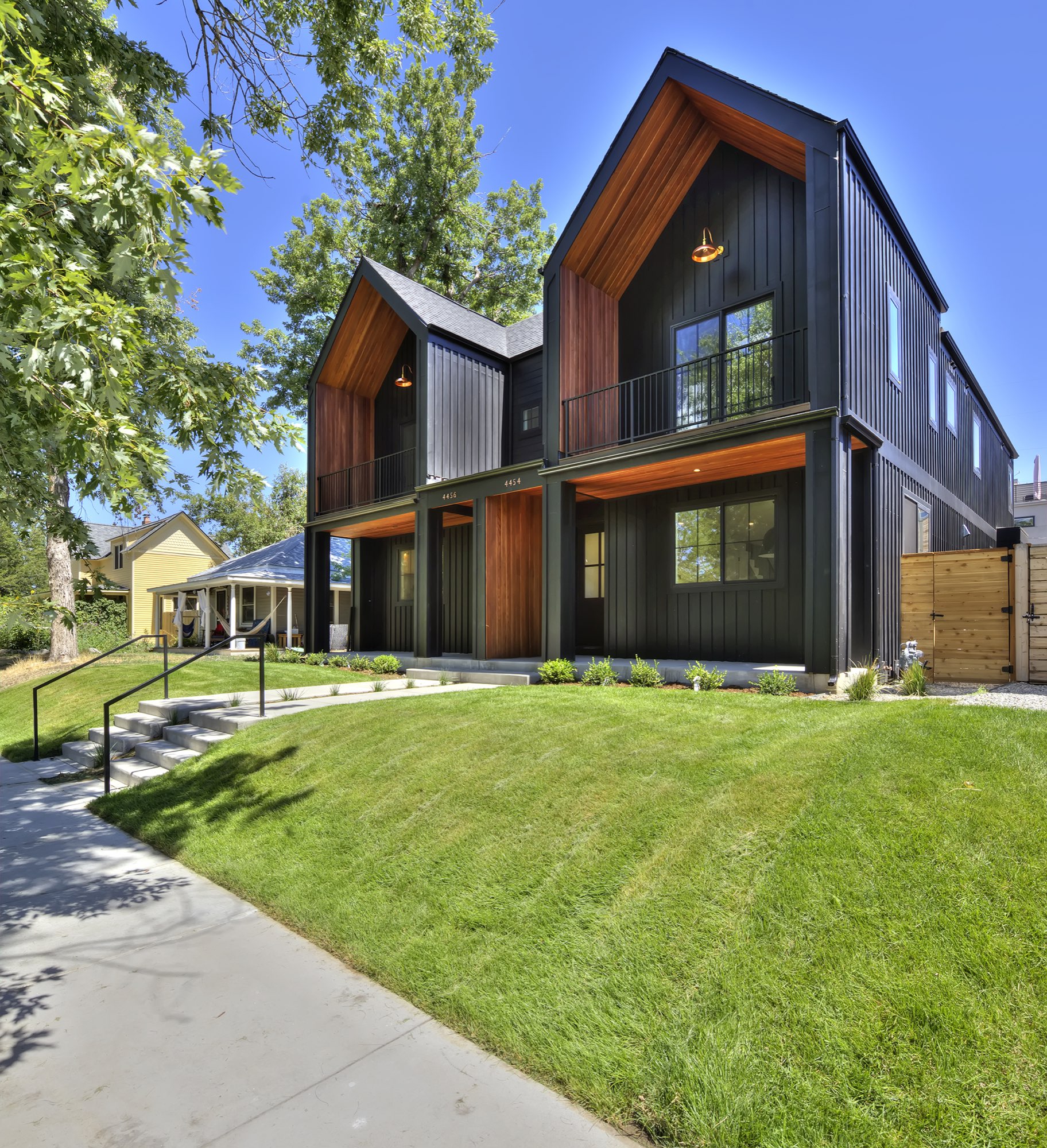 Front of the Nordic farmhouse duplex with two black gables lined in wood, balconies with black railings, covered porches and a green lawn