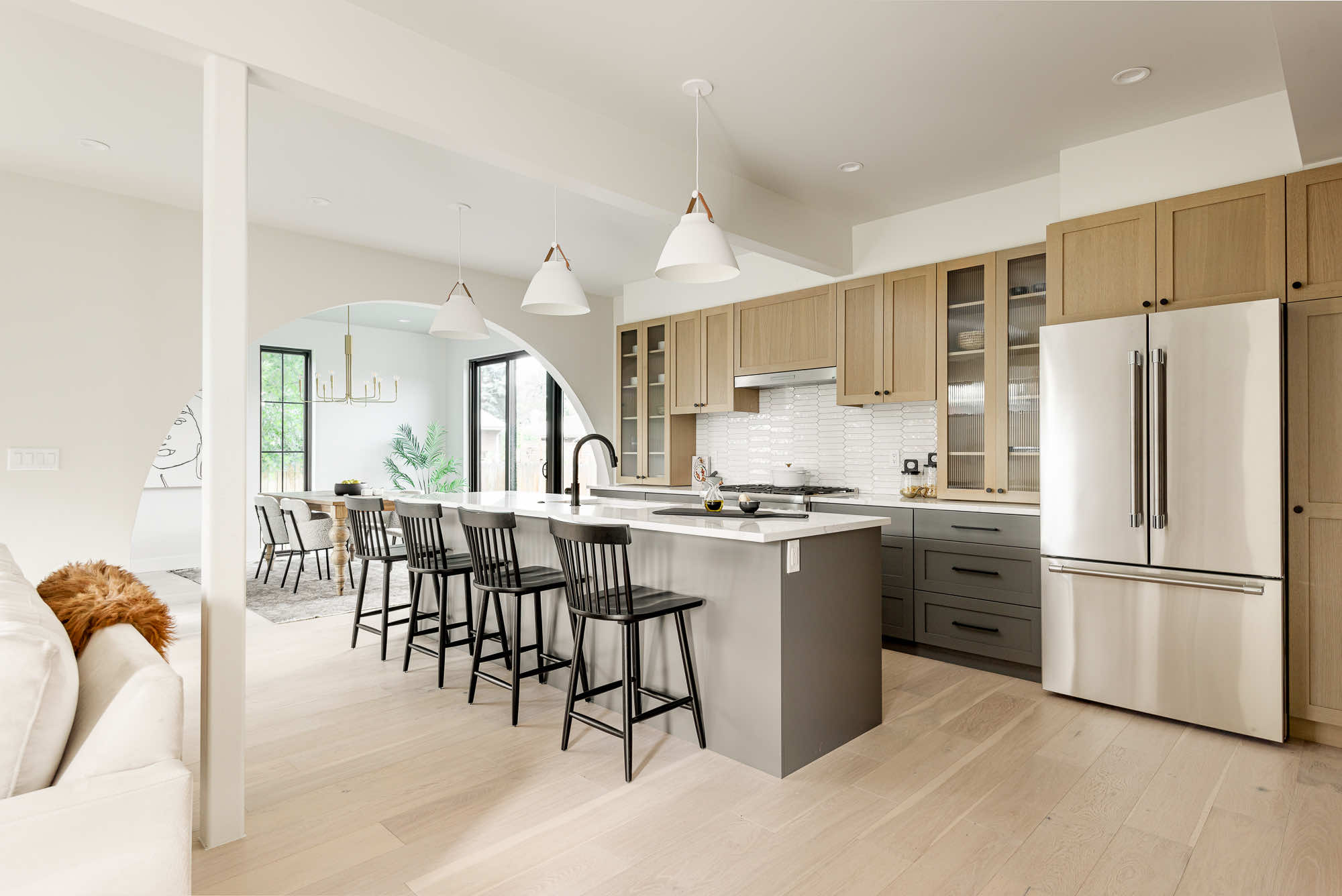 Open kitchen with a long gray island, black stools, white pendants, light wood cabinets and a round arch to the dining room