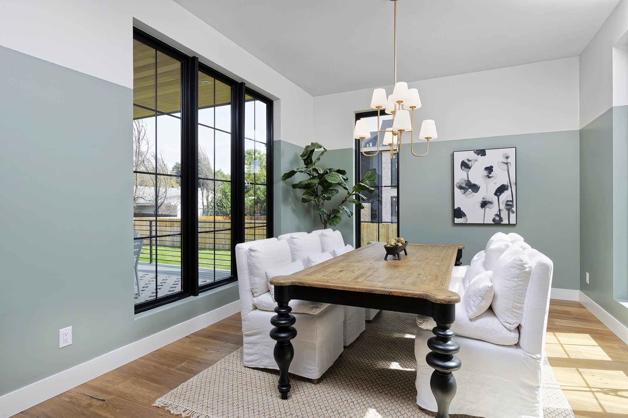 Dining room with sage green lower walls, tall black-framed windows, a brass chandelier and a wood table