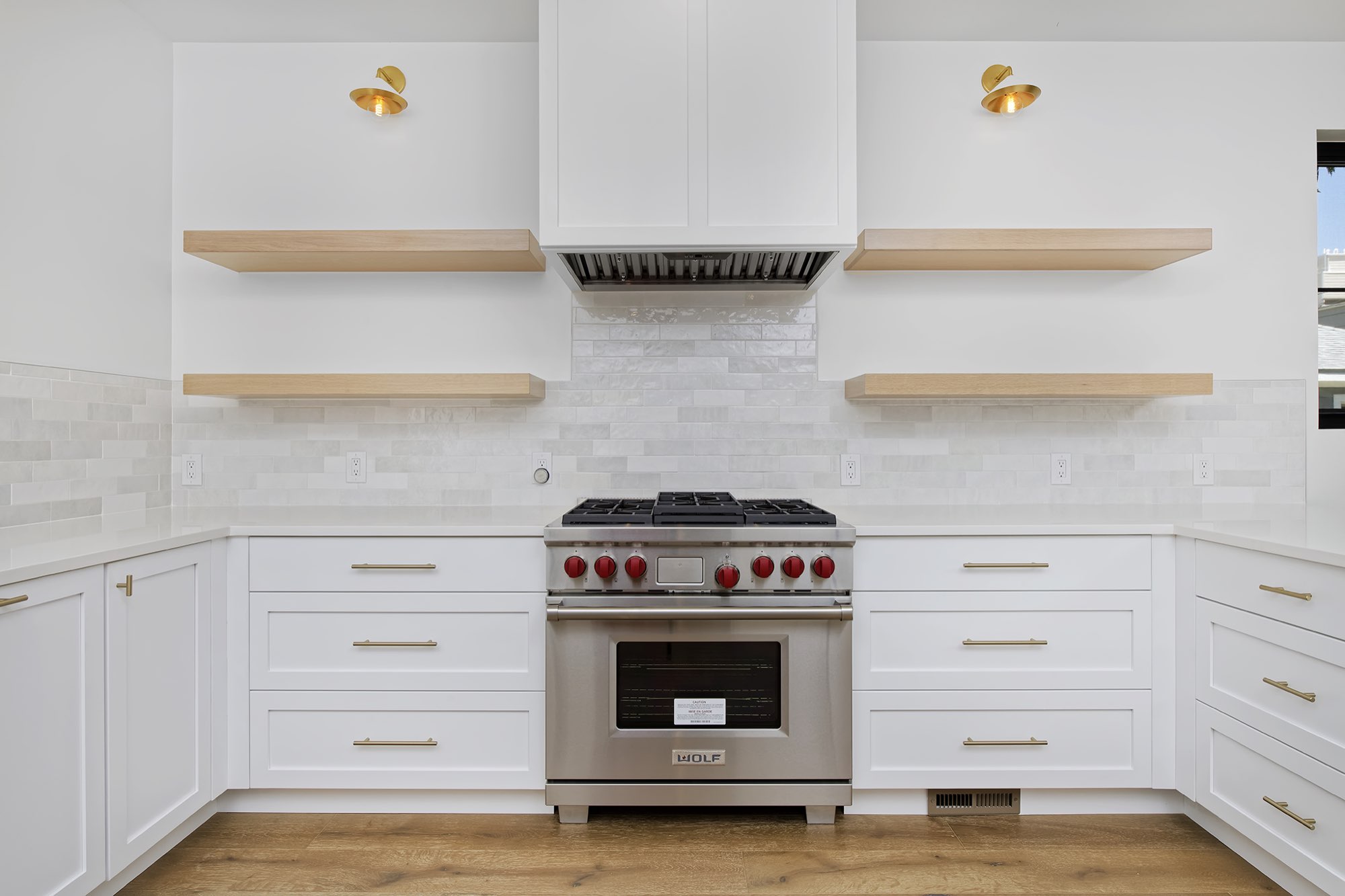Stainless range under a white hood with floating wood shelves, brass sconces and stone-look tile