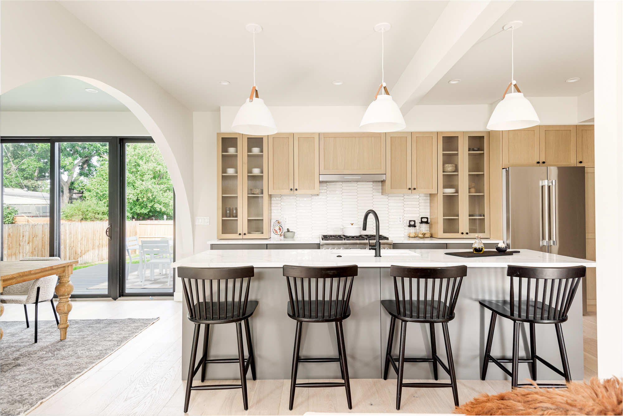 Kitchen island with four black stools, light wood cabinets with glass doors and a white tile backsplash