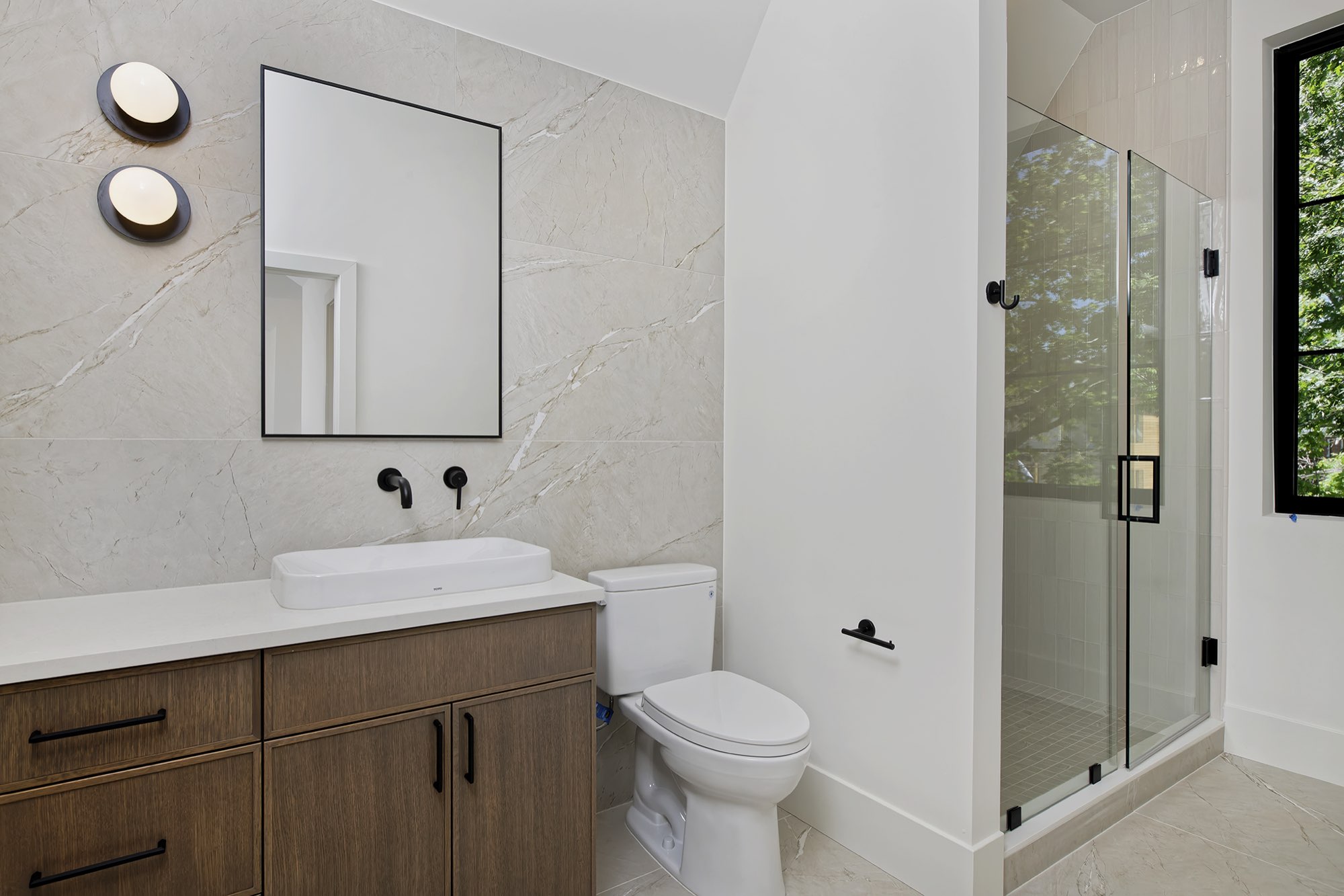 Bathroom with gray veined tile wall, floating wood vanity, black wall faucet and a glass shower