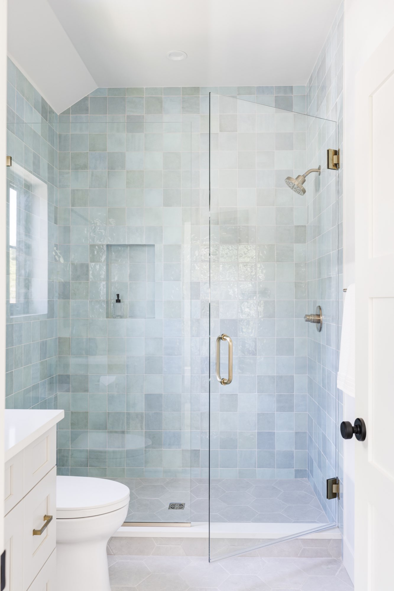 Guest shower in soft blue-green tile with a frameless glass door, brass handle and brass shower fixtures