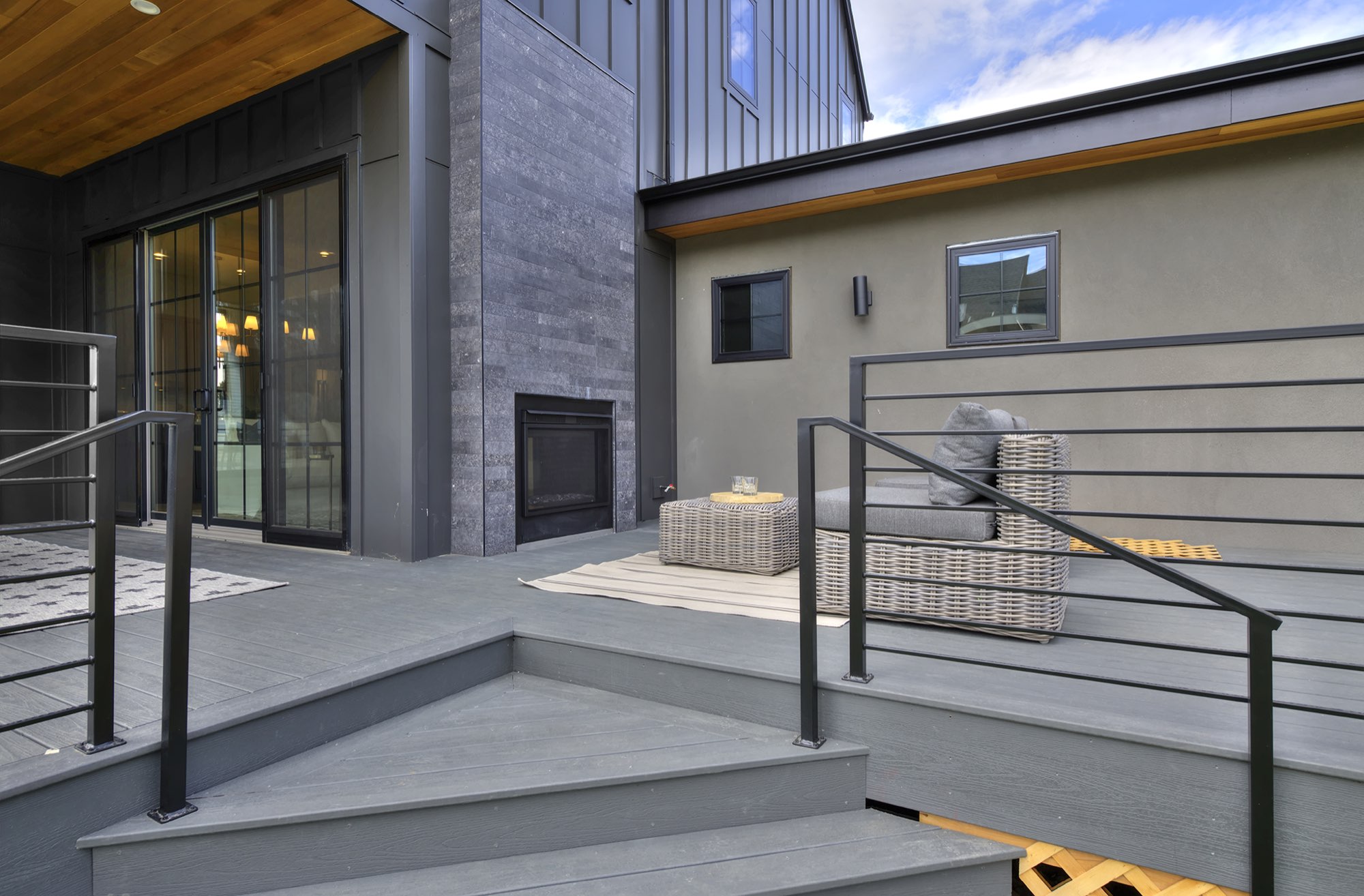 Deck steps and black railing beside the outdoor fireplace and sliding doors, with a wicker chair on the gray deck boards