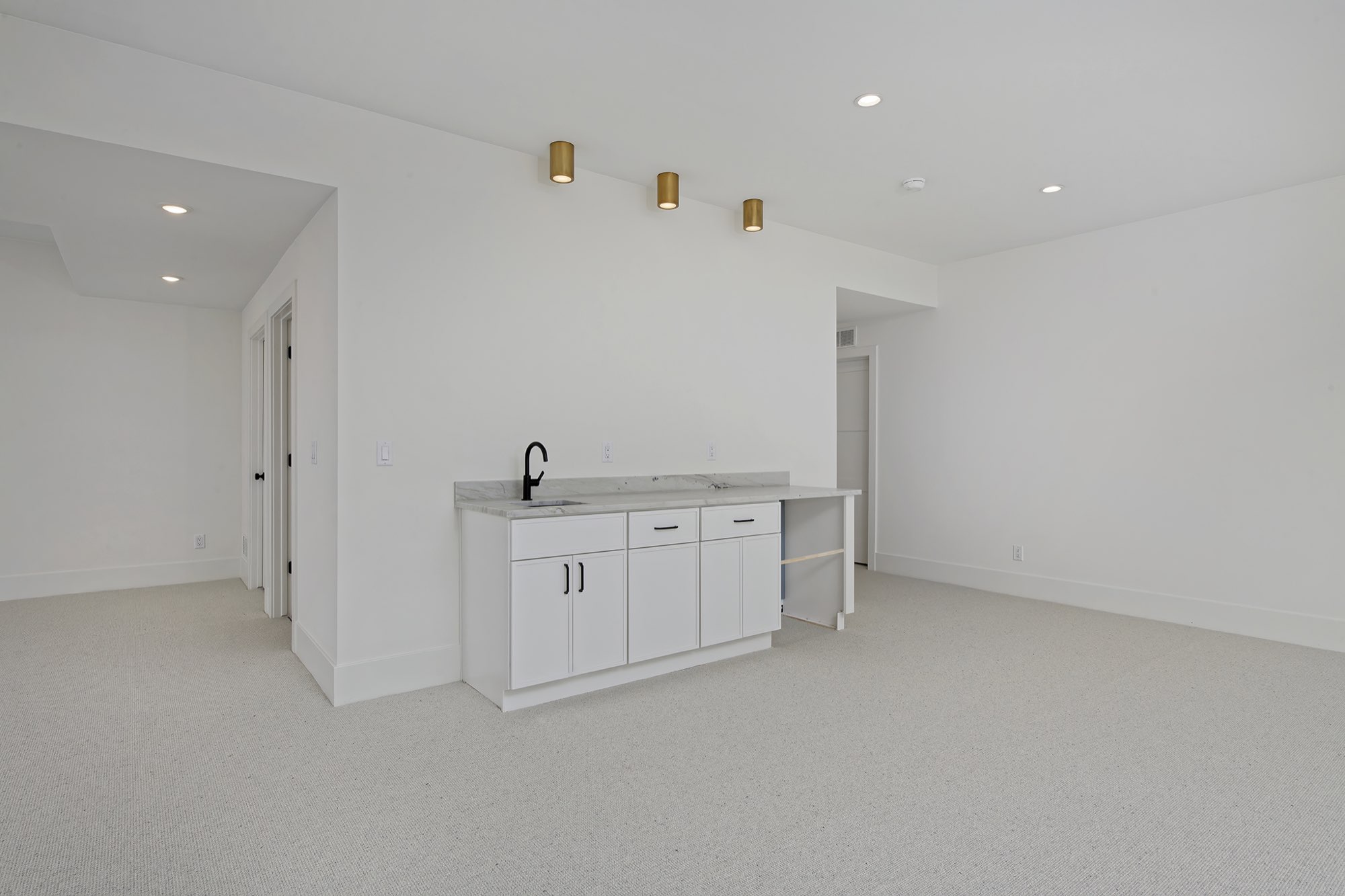 Lower level open room with carpet, brass ceiling lights and a wet bar with white cabinets and a black faucet