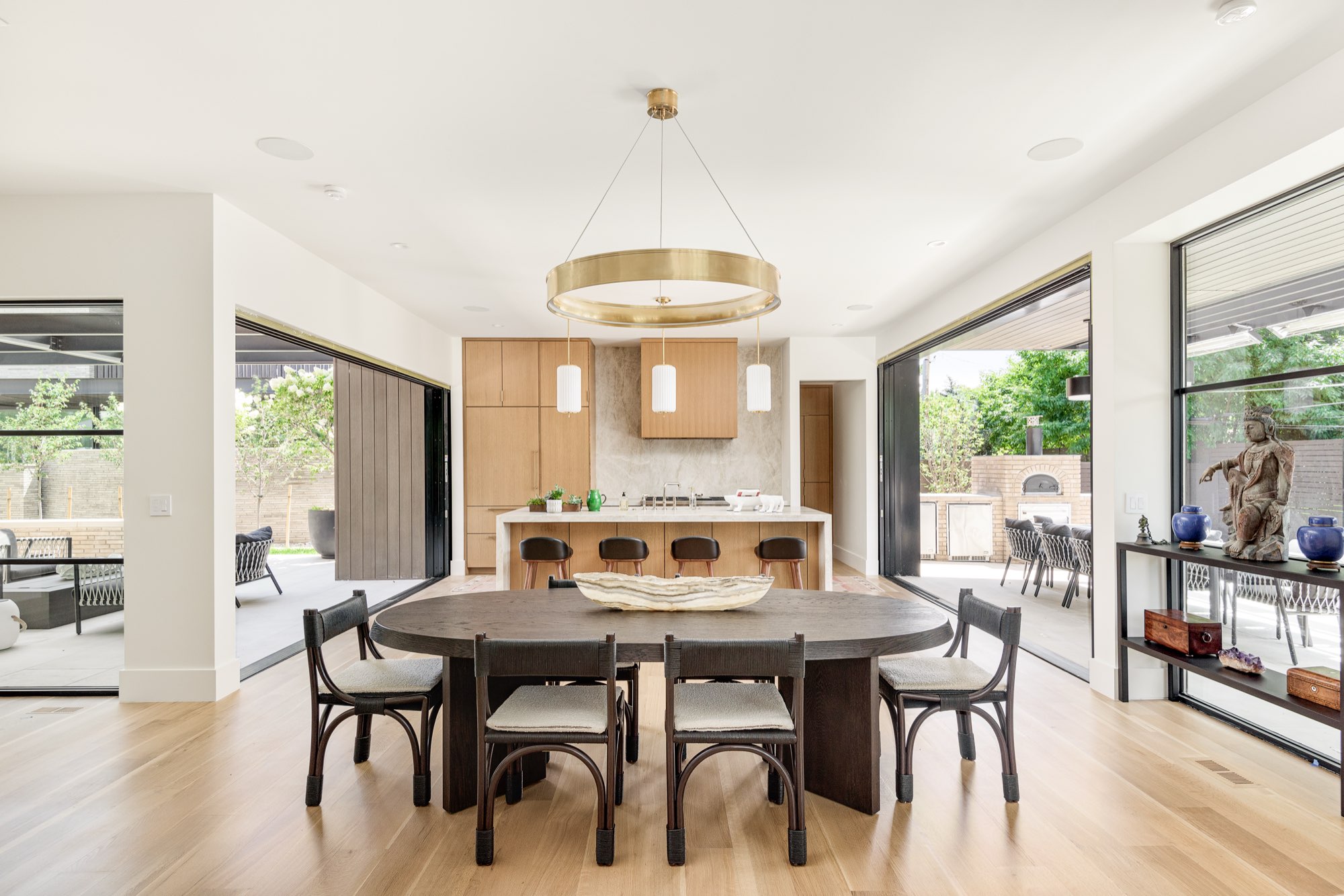 Dining table in front of the kitchen island under a brass ring pendant, with glass walls toward the patio