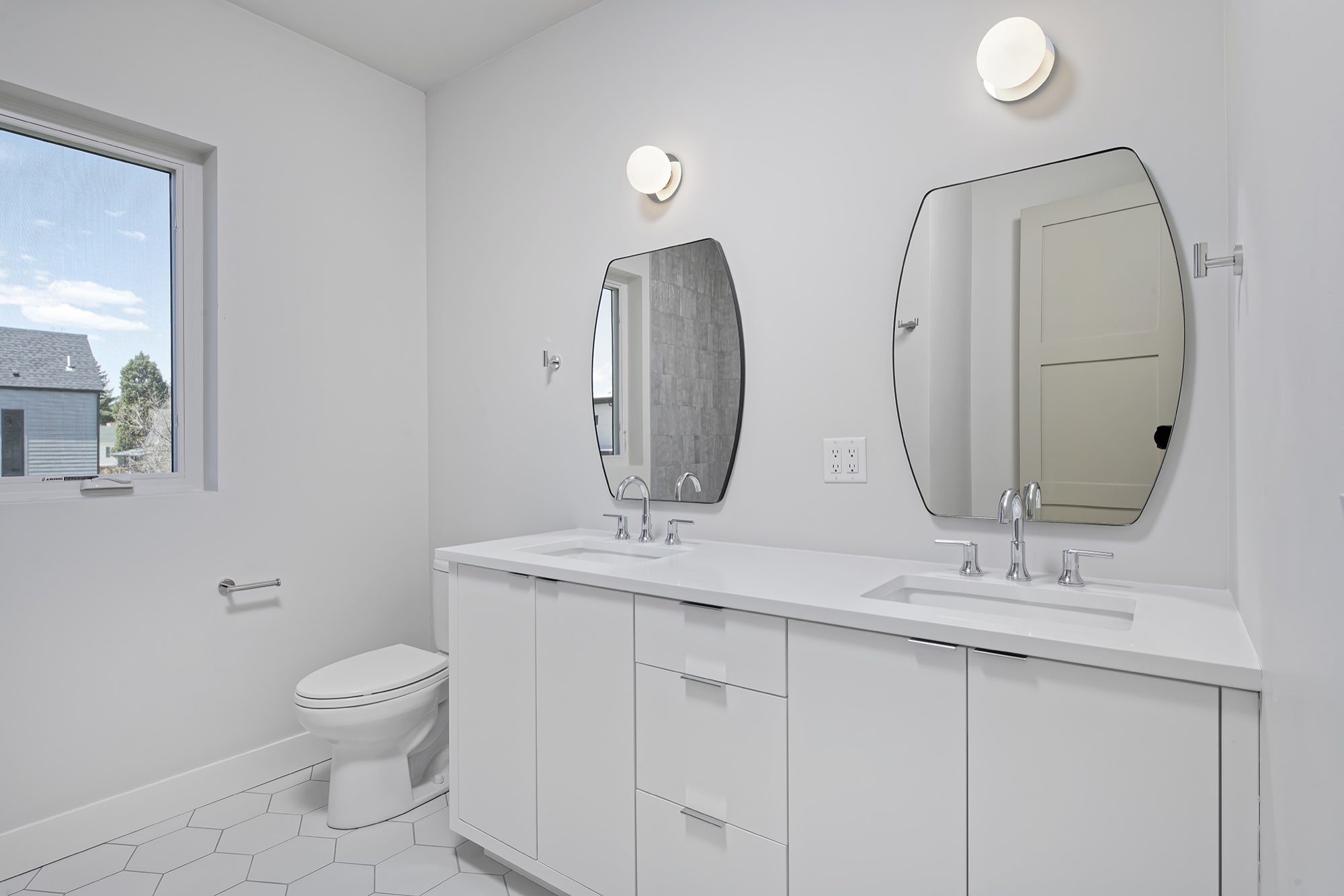 Hall bath with a white double vanity, two black-framed shaped mirrors, hex tile floor and a window