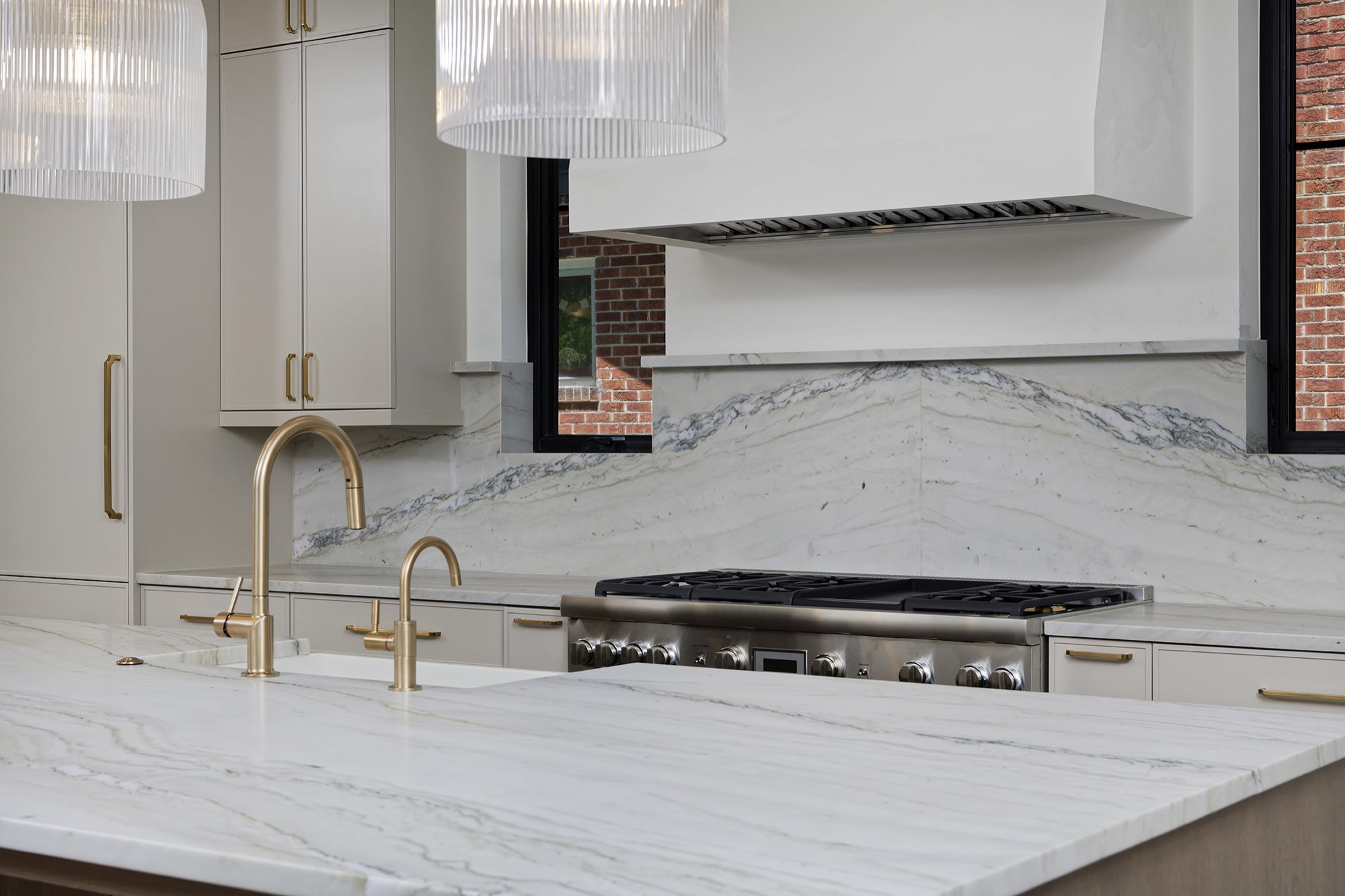 Brass faucets, white veined stone counter and backsplash, and a gas range under a white hood
