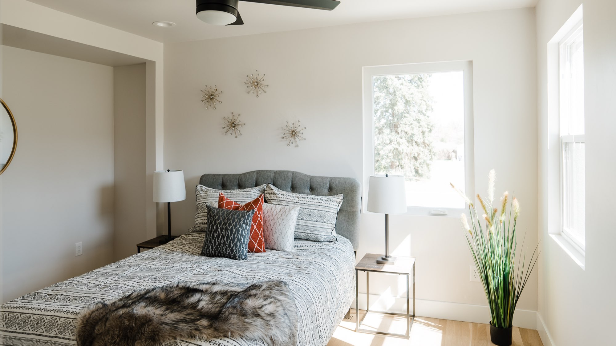 Bedroom with a tufted gray headboard, patterned bedding, two lamps, a ceiling fan and windows on two walls