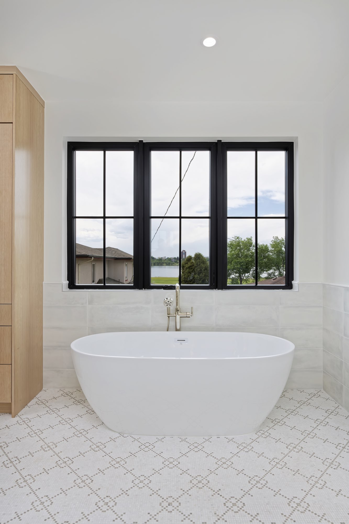 Freestanding soaking tub in front of three black-framed windows with a view toward the lake
