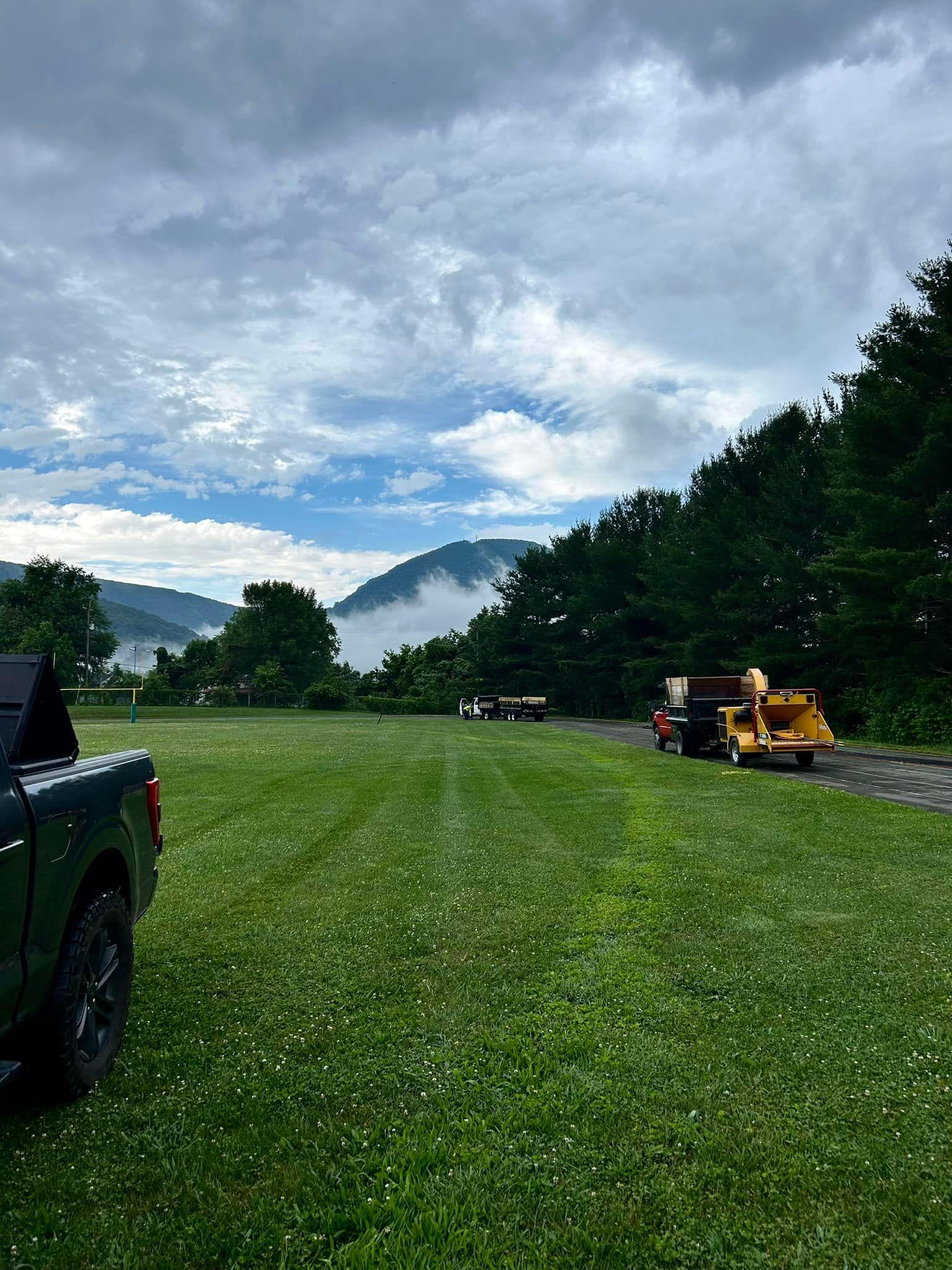 NRV Tree Pro trucks, dump trailers and a wood chipper staged along an access track beside a wide mown field, with the New River Valley mountains behind